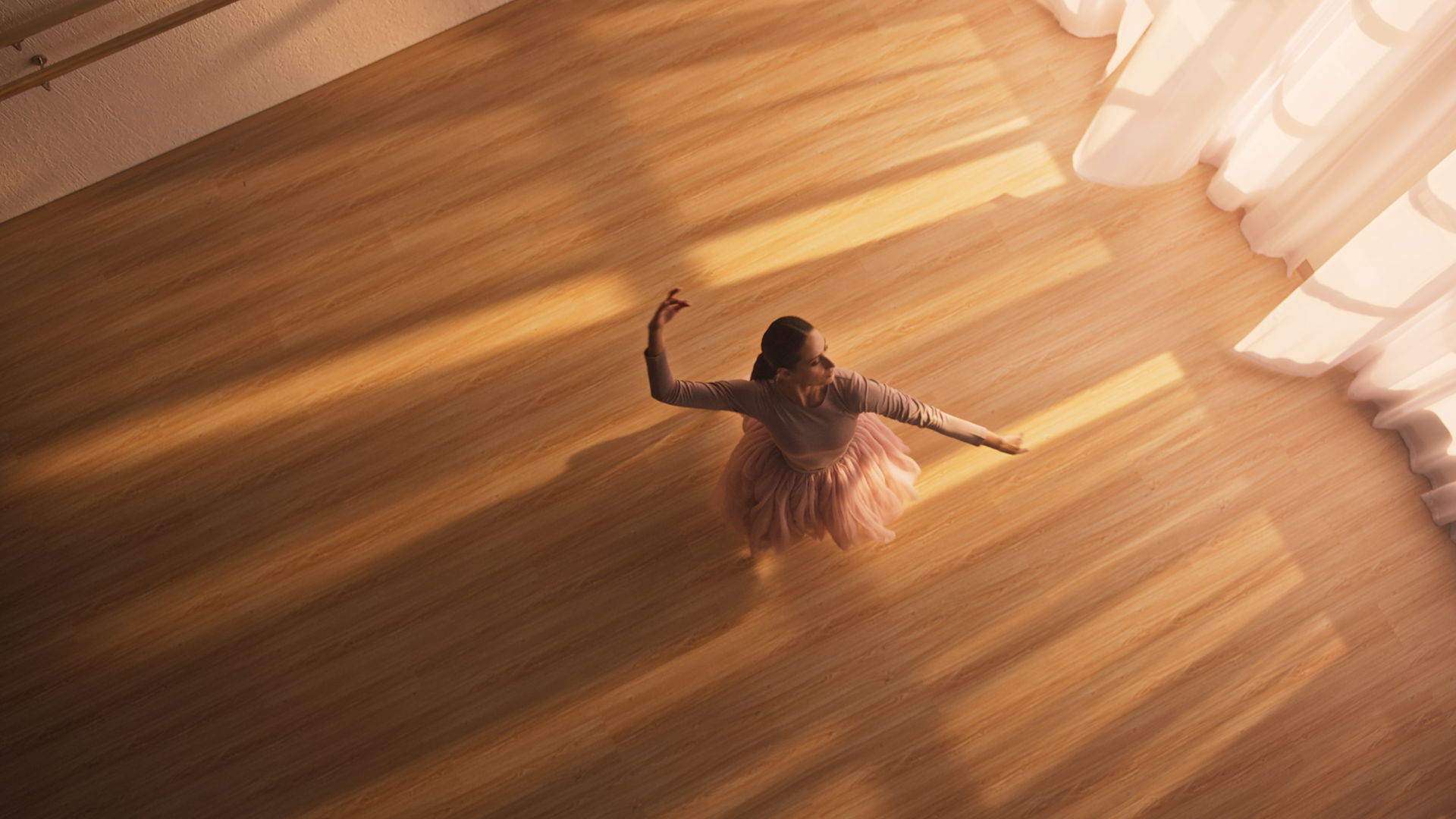 A ballerina in a pink tutu dances alone in a sunlit studio, seen from above. Warm light streams through large windows, casting shadows on the wooden floor.