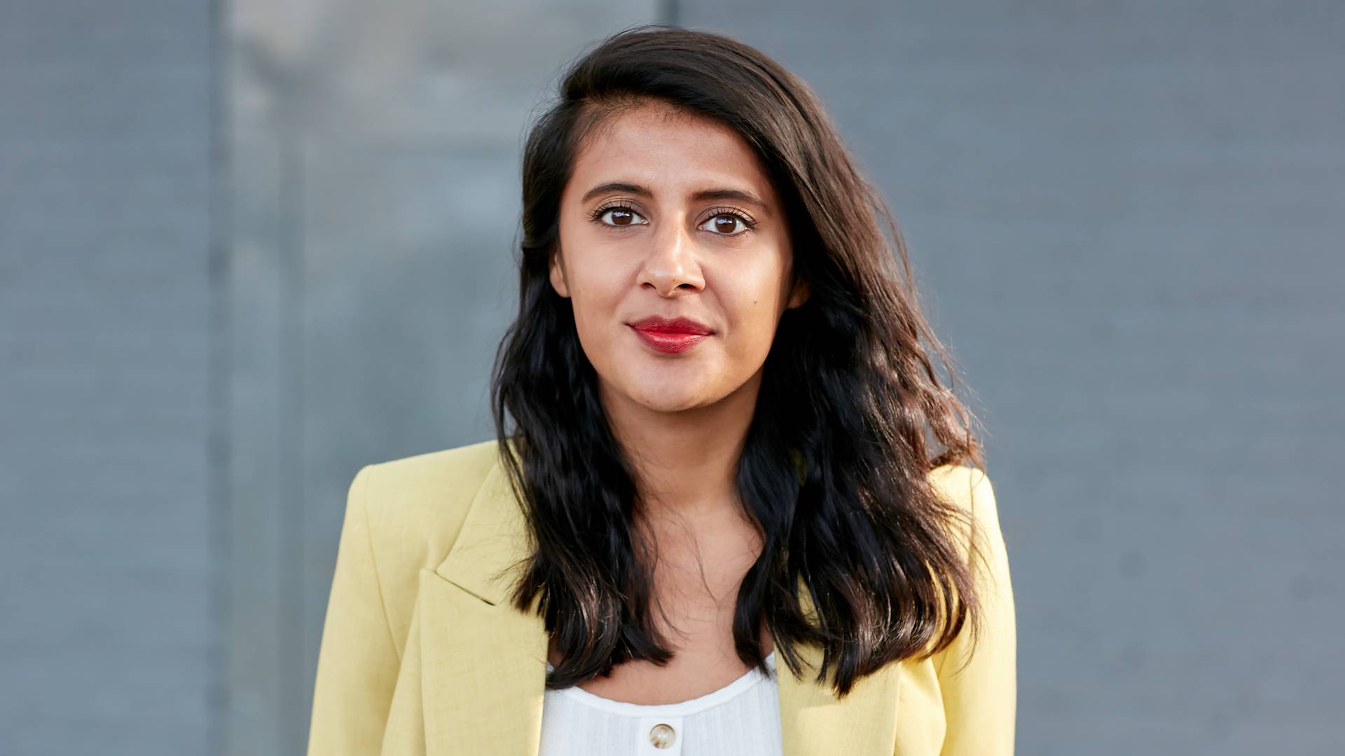 A woman with long dark hair, wearing a pale yellow blazer over a white top, stands in front of a plain gray background, looking directly at the camera with a neutral expression.