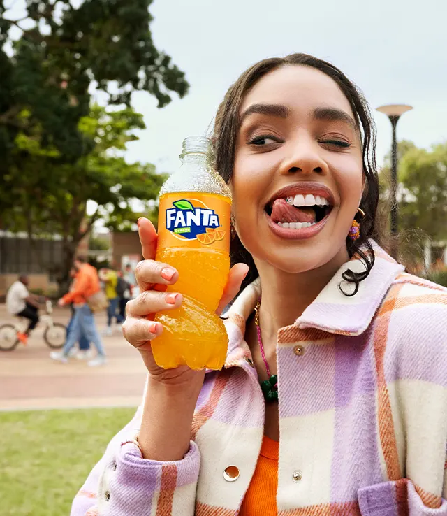 A young woman outdoors holds a bottle of orange Fanta, smiling playfully with her tongue out. She wears a checked jacket and stands in a park with people in the blurred background.