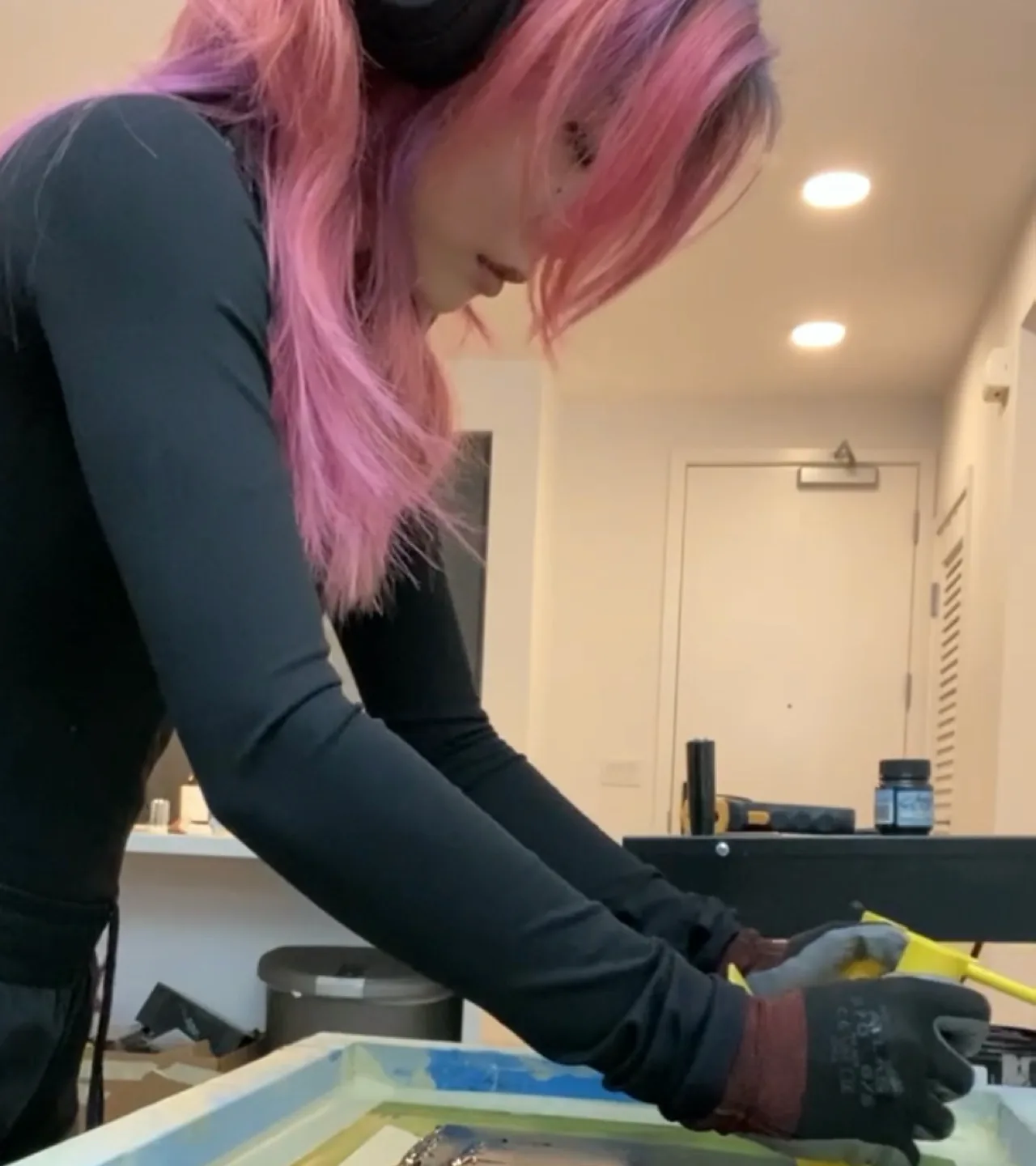 A person with long pink hair, wearing headphones and gloves, is focused on working with a screen printing frame in a well-lit room with white walls.