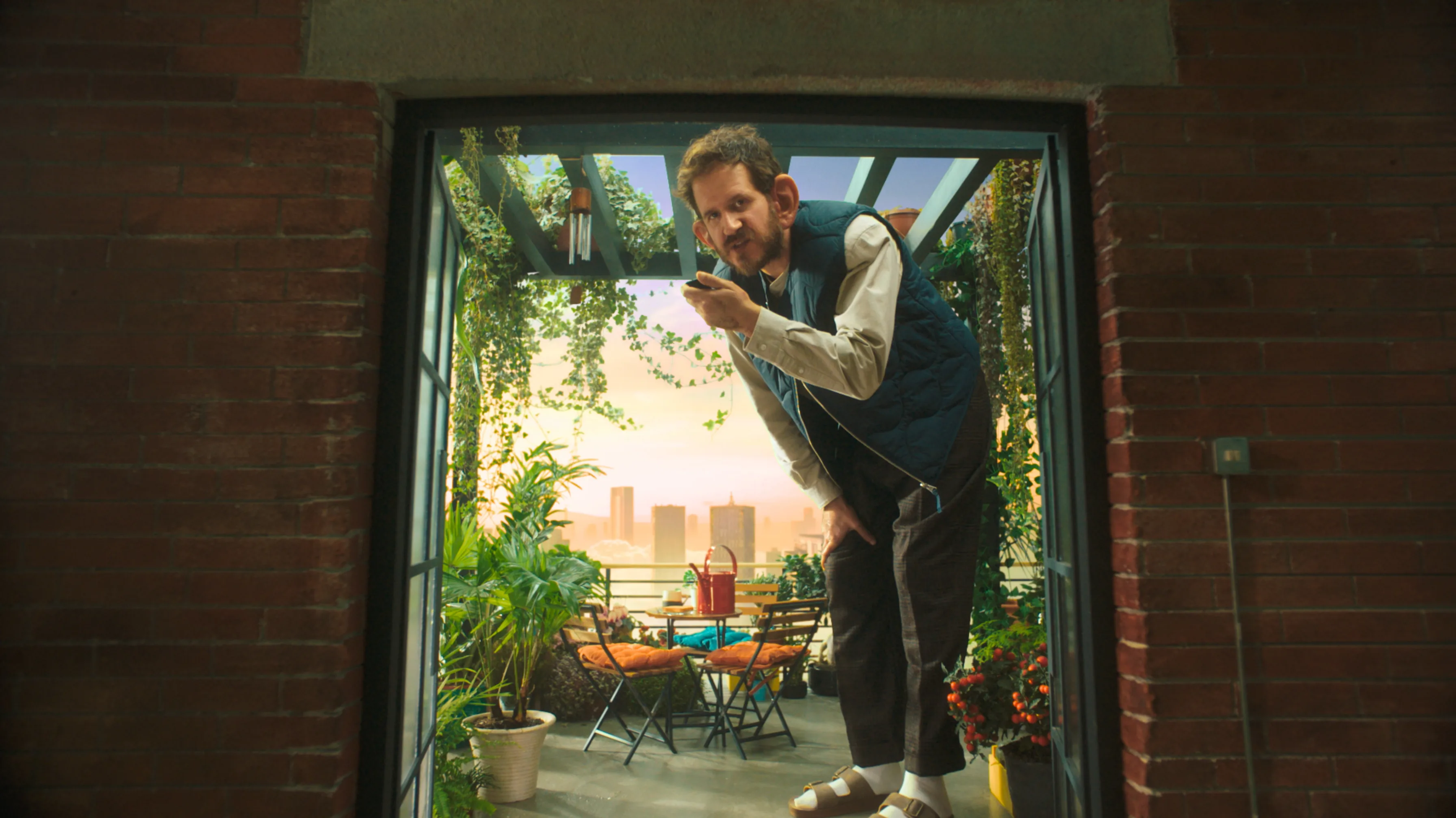 A man in sandals and a vest leans through a doorway, holding a phone. Behind him is a balcony with lush plants, outdoor furniture, and a cityscape at sunset in the background.