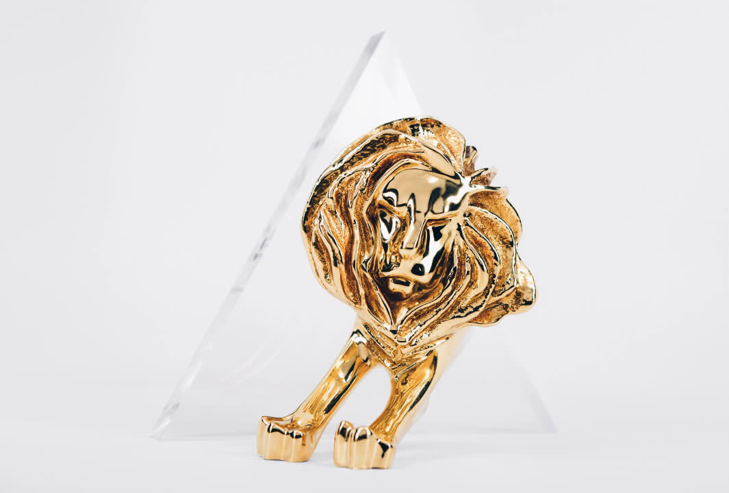 A gold lion trophy with a detailed mane stands in front of a clear, triangular glass base against a plain white background.