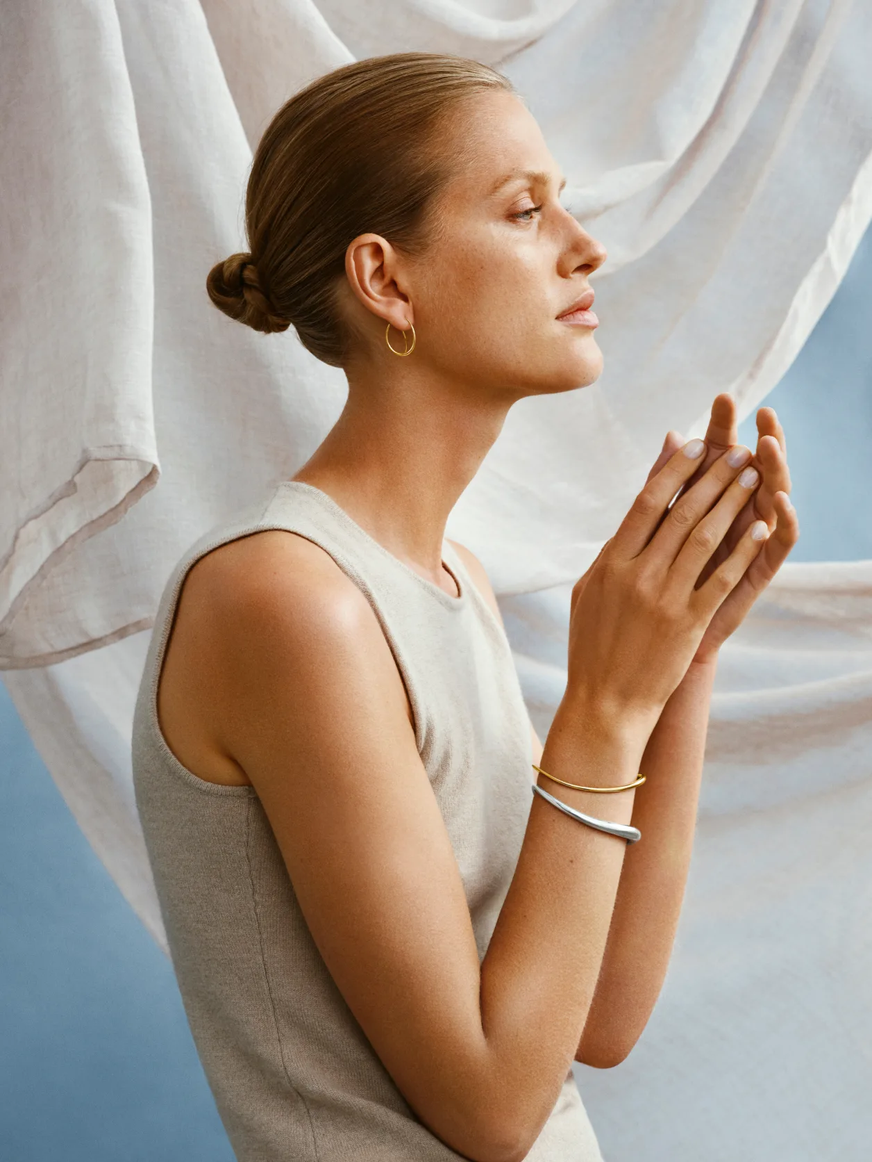 A woman with light brown hair in a sleek bun poses in profile against flowing white fabric. She wears a sleeveless beige top, gold hoop earrings, a bracelet, and holds her hands near her face in a thoughtful gesture.