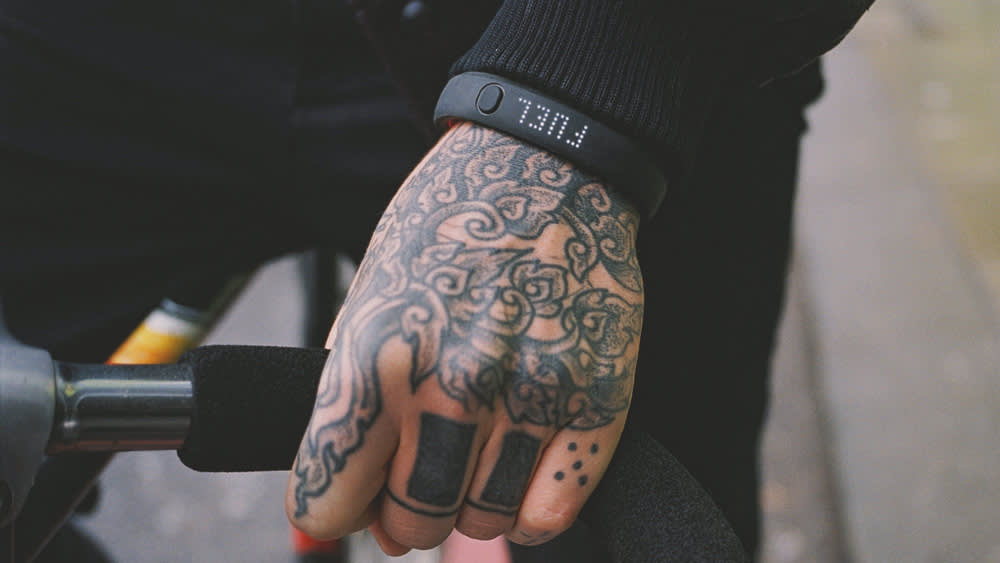 A close-up of a tattooed hand gripping a bicycle handlebar. The person is wearing a black wristband with the word FULL printed on it. The background is blurred.