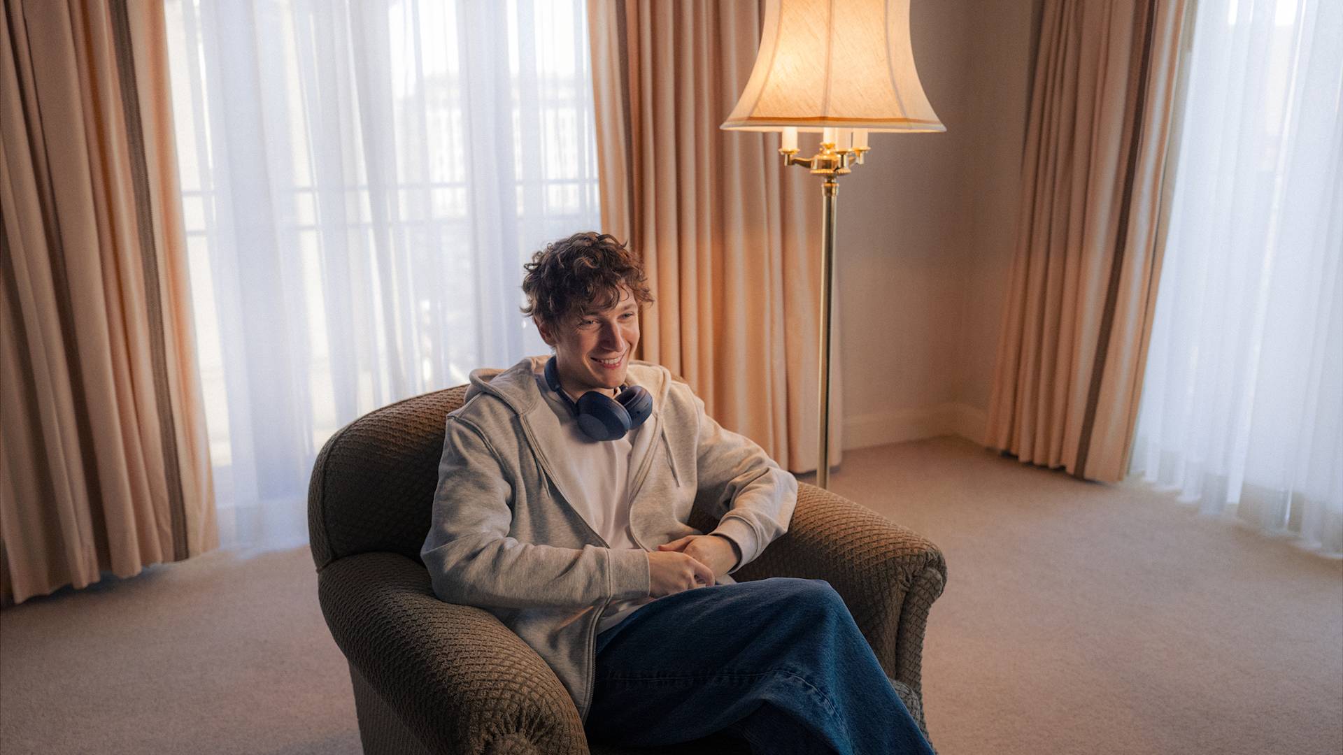 A person with curly hair, wearing a gray hoodie and blue headphones around their neck, sits smiling in an armchair in a bright room with beige curtains and a floor lamp.