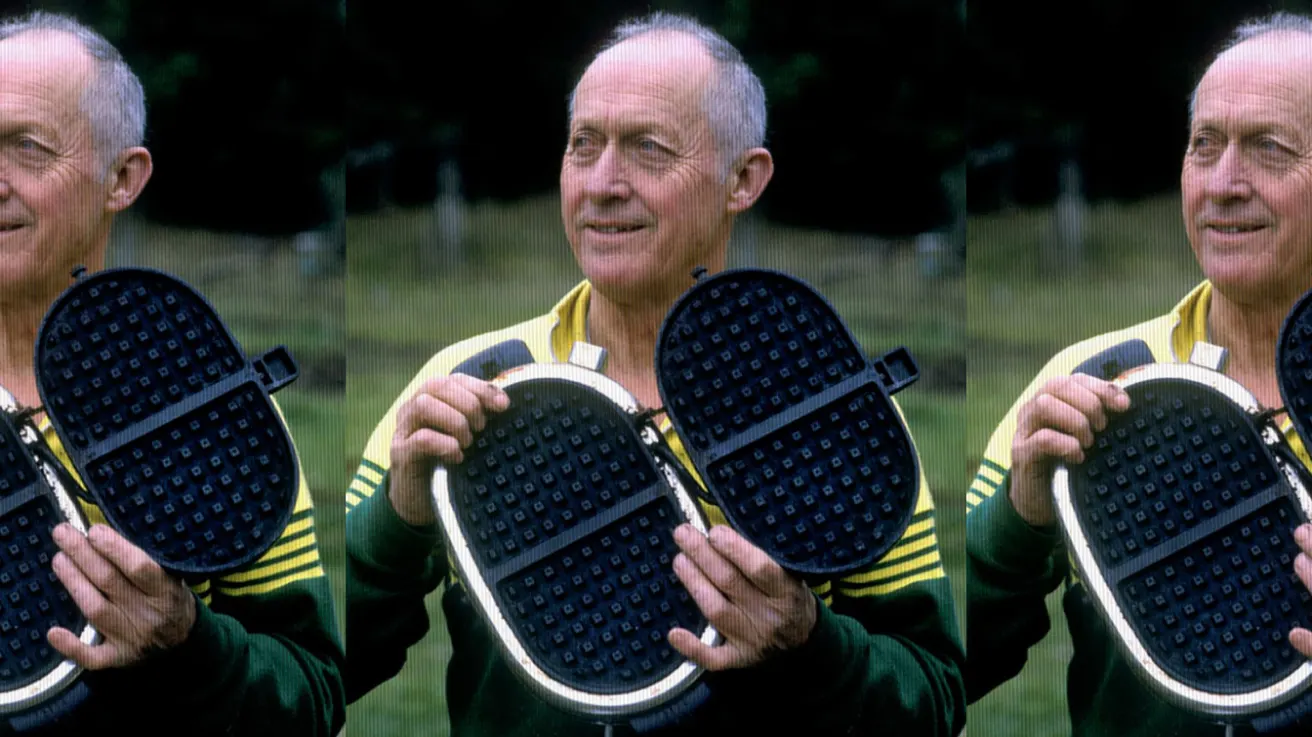 An older man in a green and yellow jacket smiles while holding open a black waffle iron outdoors, with trees blurred in the background.