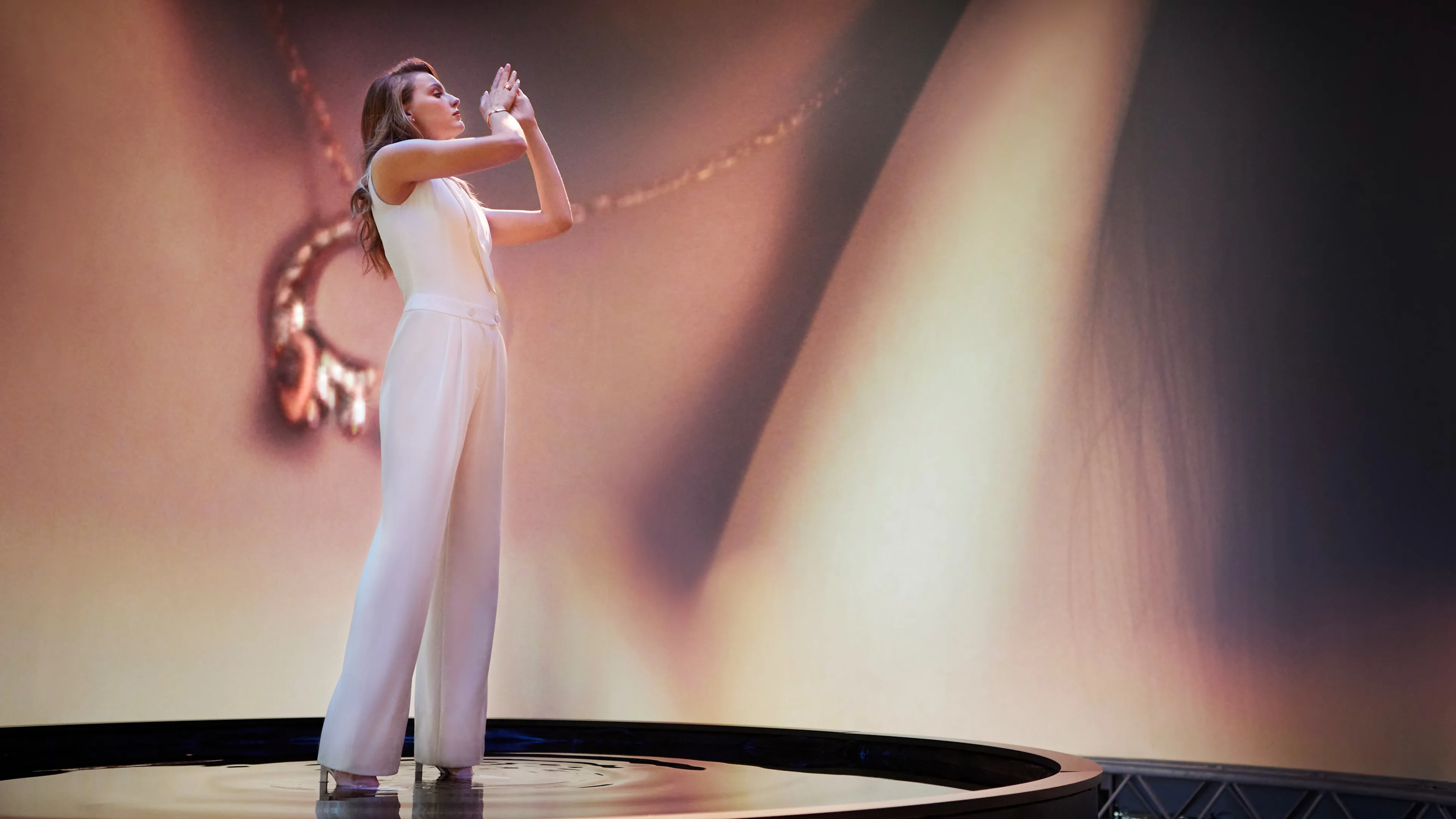 A woman in a white sleeveless jumpsuit stands on a circular stage, raising her hands gracefully. A large, blurry image of a necklace and skin is projected behind her, creating an artistic, dramatic backdrop.