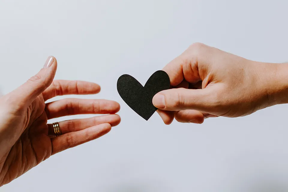 A hand extends a paper heart towards another open hand, suggesting an act of giving or sharing love and kindness. The background is plain and light-colored.