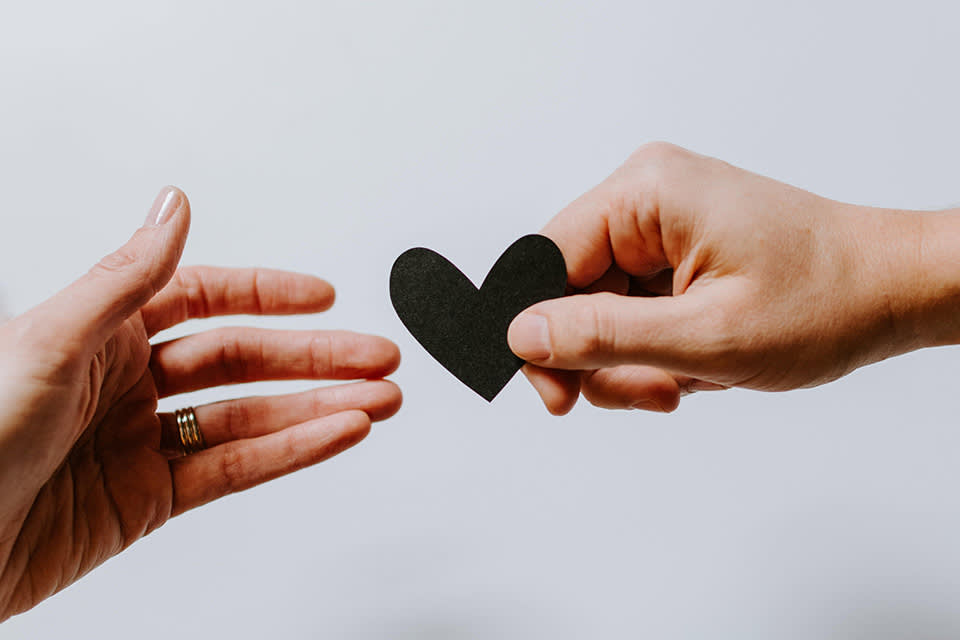 A hand extends a paper heart towards another open hand, suggesting an act of giving or sharing love and kindness. The background is plain and light-colored.