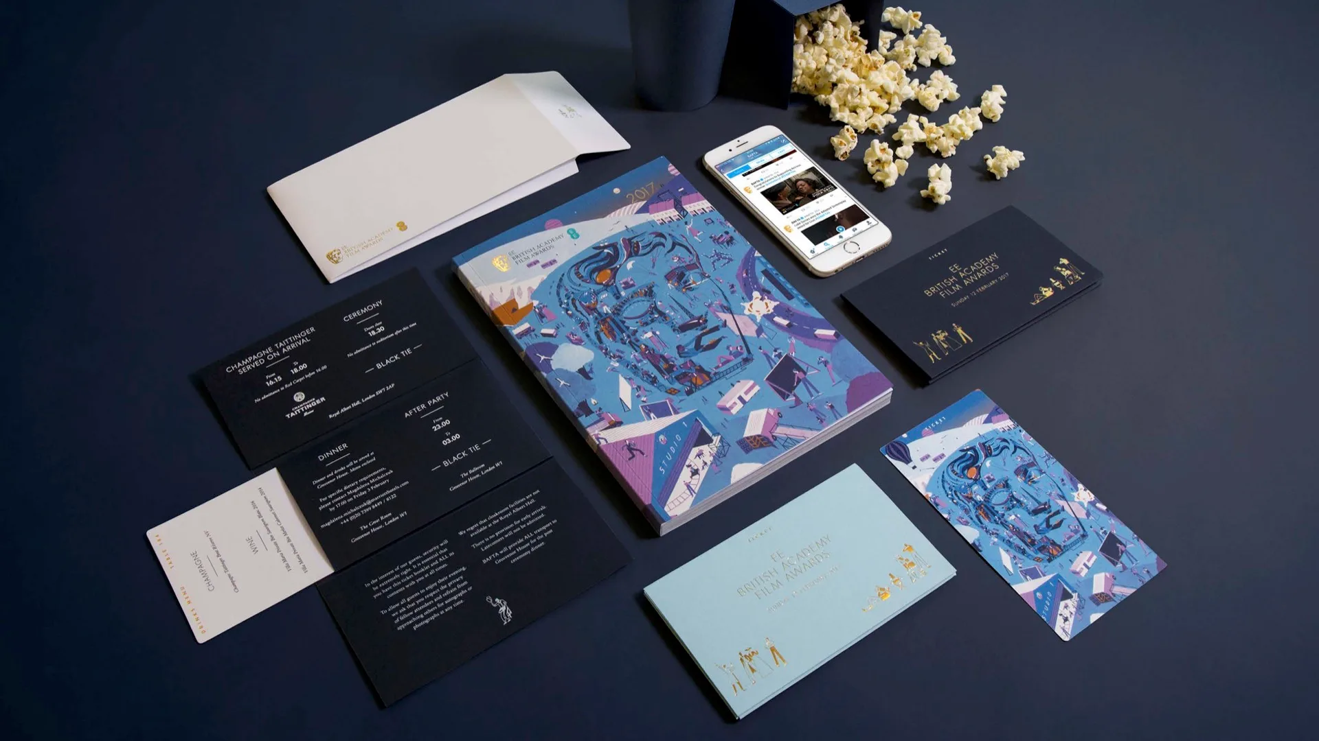 A flat lay of stationery items, invitations, illustrated booklets, a smartphone, and a tipped cup with spilled popcorn on a dark blue surface. The booklets feature vibrant illustrations of a face and geometric shapes.