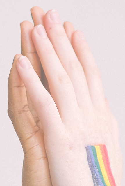 Two hands of different skin tones are pressed together, with one hand showing a rainbow flag painted on it, symbolizing LGBTQ+ pride and unity.