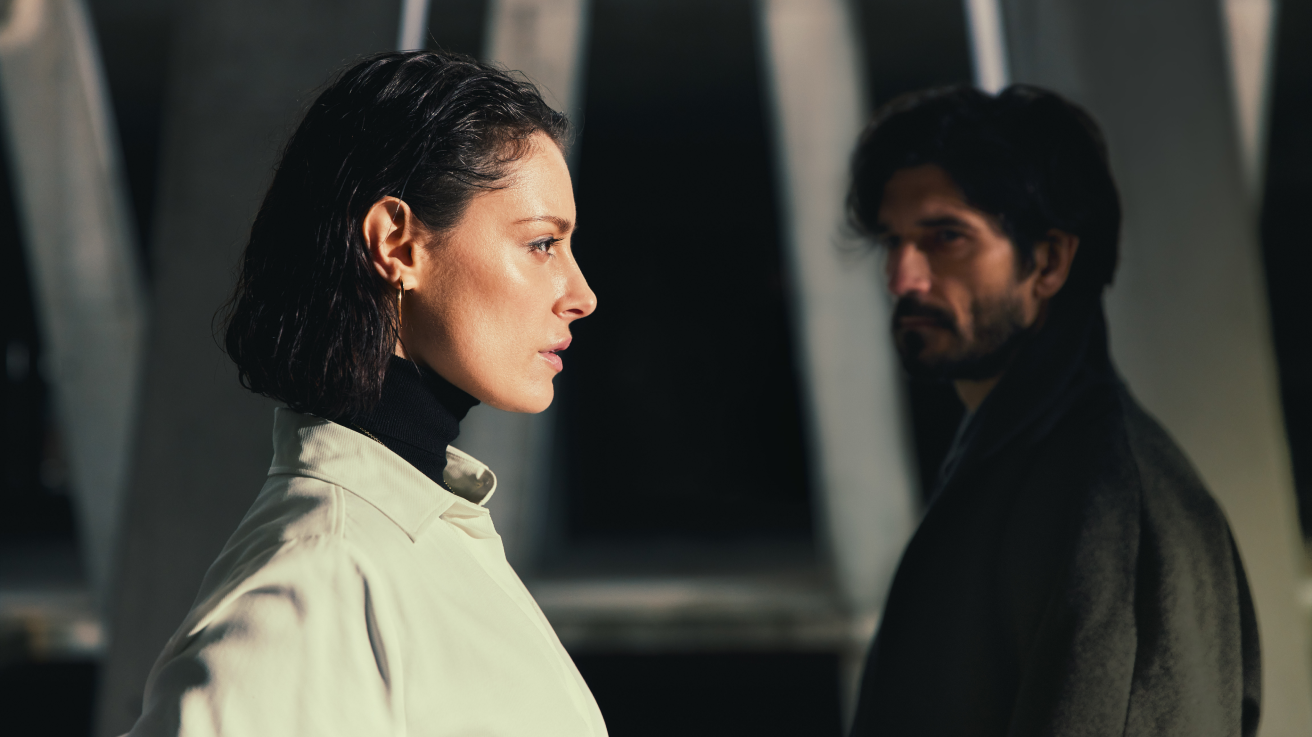 A woman in a light coat looks ahead with a serious expression, while a man in dark clothing stands behind her in shadow, both positioned against a background of tall, blurred architectural columns.