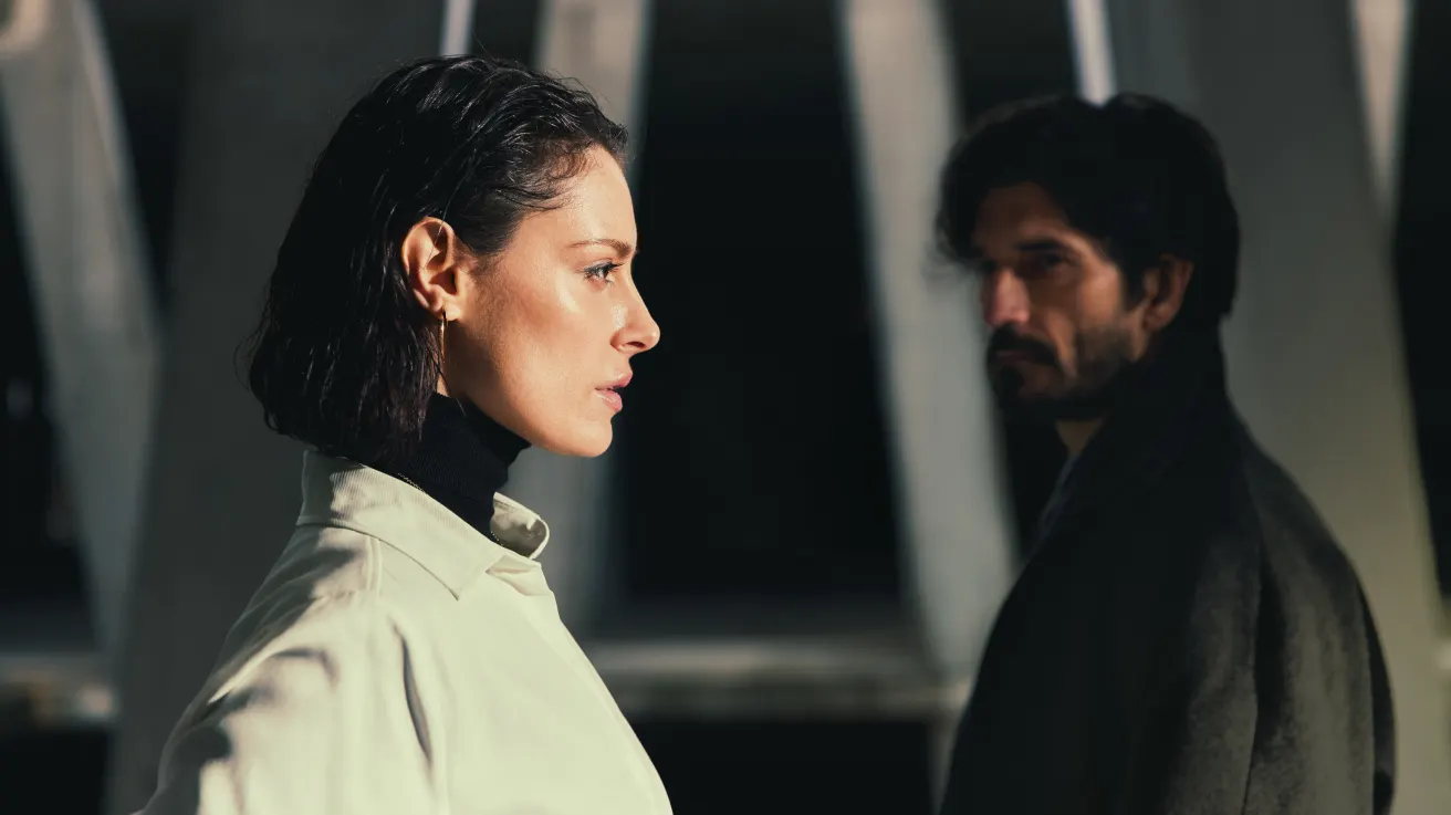 A woman in a light coat looks ahead with a serious expression, while a man in dark clothing stands behind her in shadow, both positioned against a background of tall, blurred architectural columns.