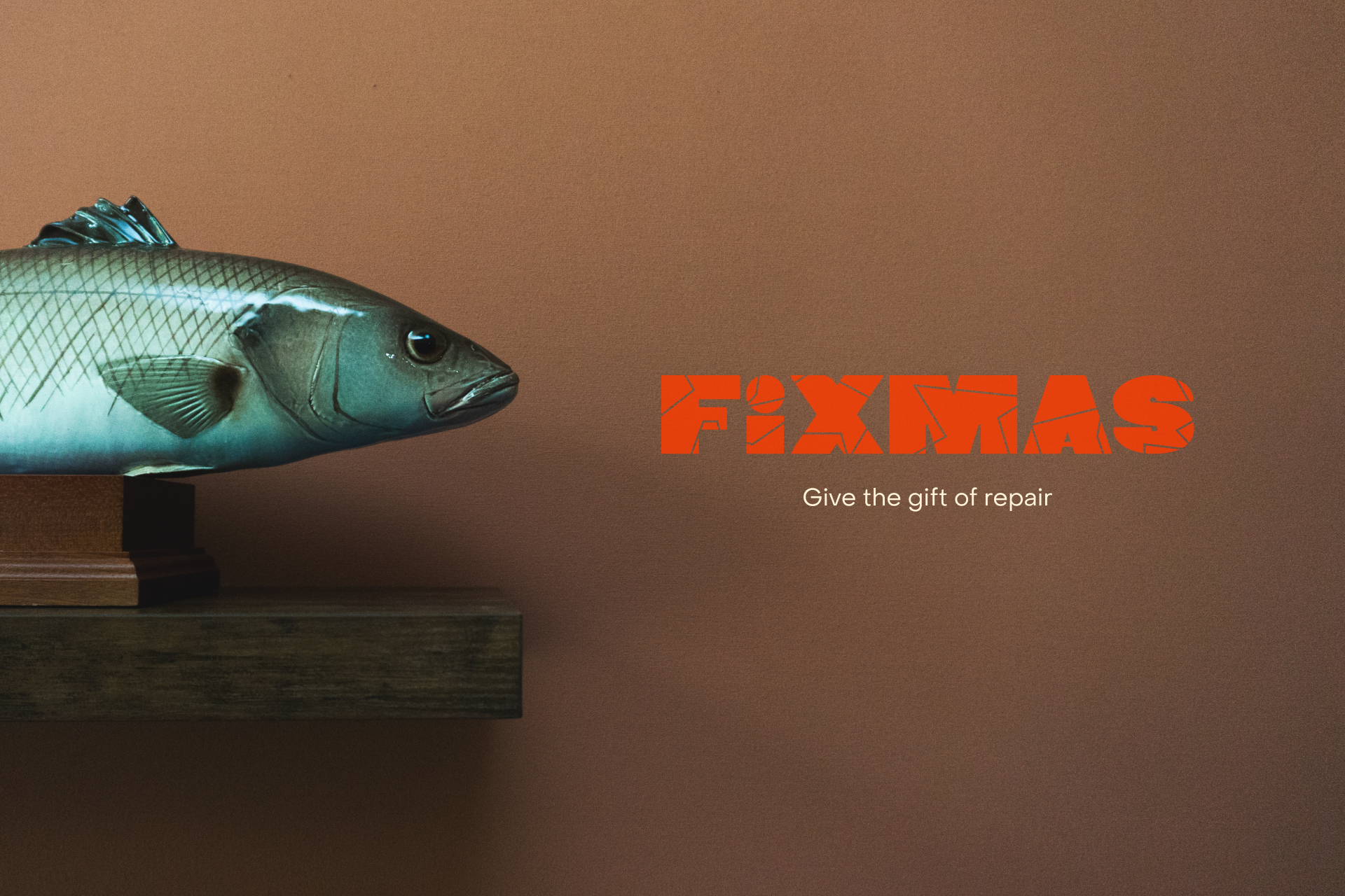 A mounted fish on a wooden shelf against a brown wall. To the right, bold orange text reads FIXMAS with smaller white text below: Give the gift of repair.