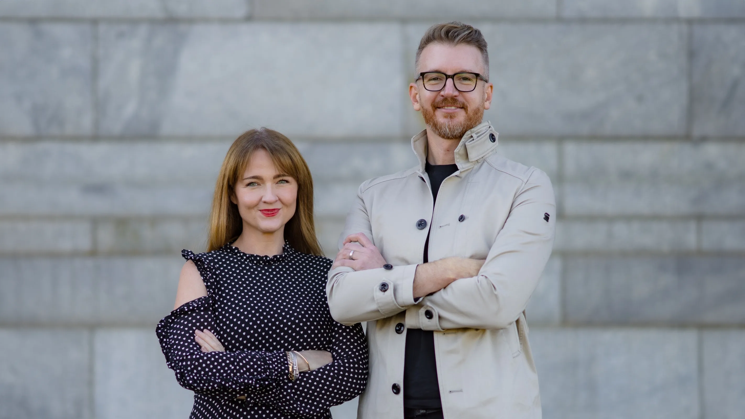 A woman in a black polka dot blouse and a man in glasses and a beige jacket stand side by side, both smiling with their arms crossed, in front of a gray stone wall.