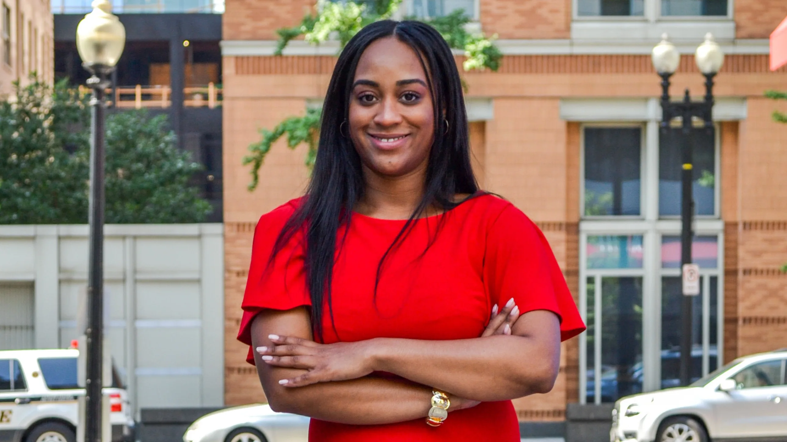A woman with long black hair, wearing a bright red dress, stands outdoors with her arms crossed, smiling confidently. Behind her are buildings, trees, and parked cars on a city street.