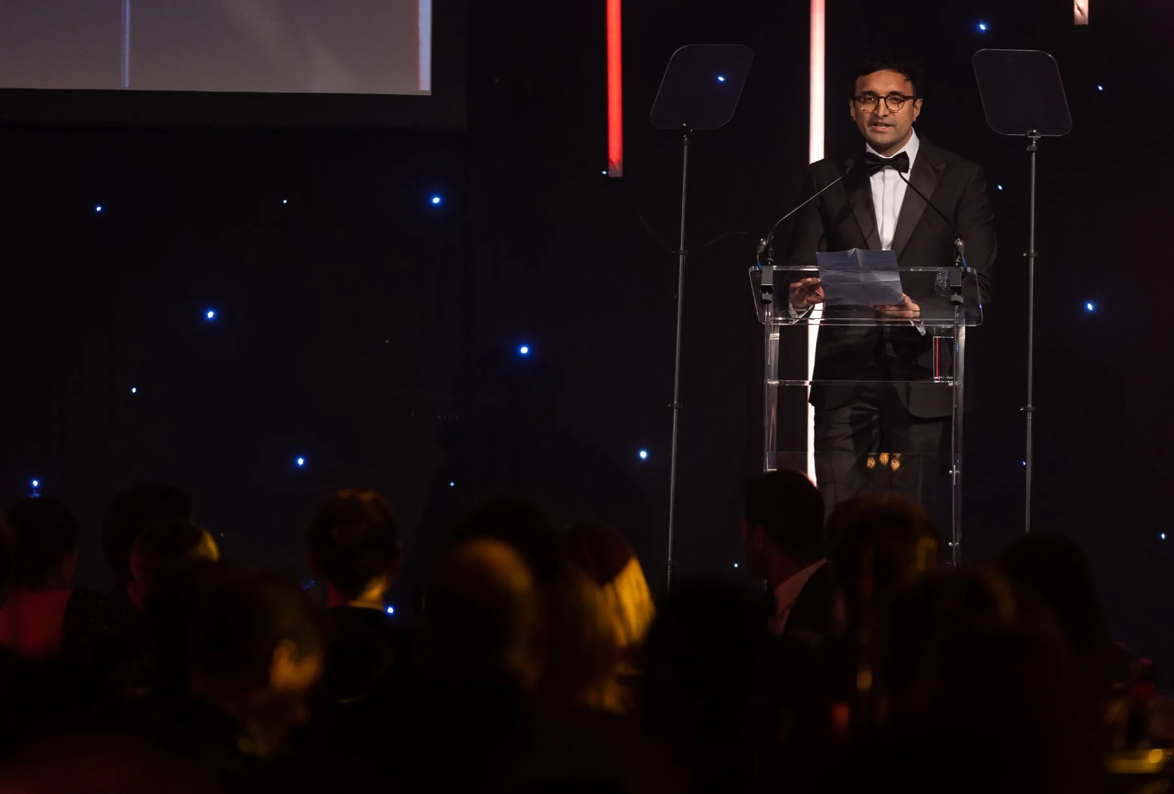 A man in a tuxedo stands at a clear podium, speaking into microphones on a dimly lit stage with blue and red lights. An audience sits in the foreground, watching him.
