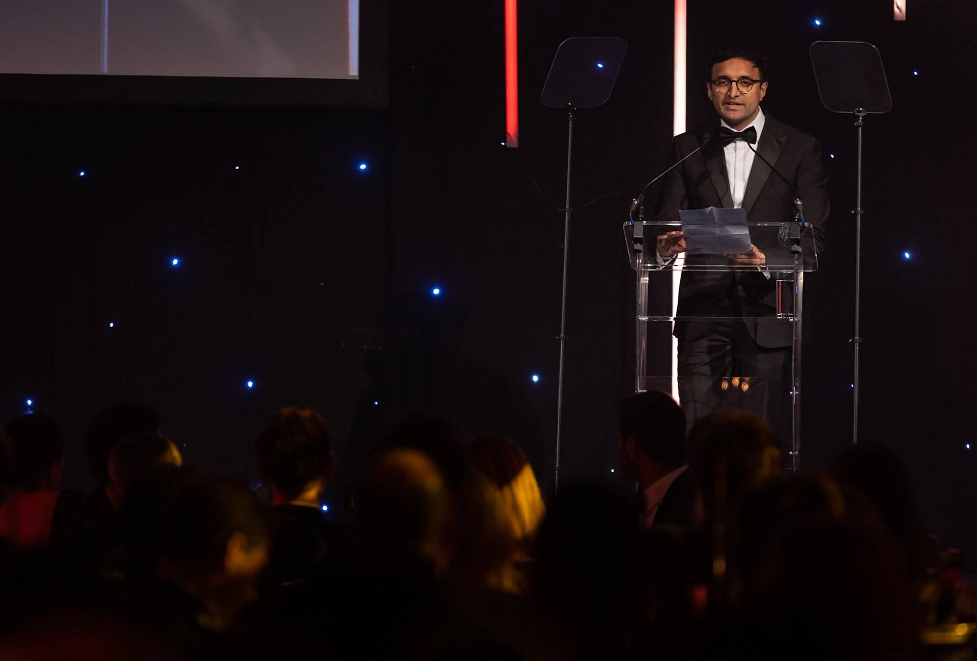 A man in a tuxedo stands at a clear podium, speaking into microphones on a dimly lit stage with blue and red lights. An audience sits in the foreground, watching him.