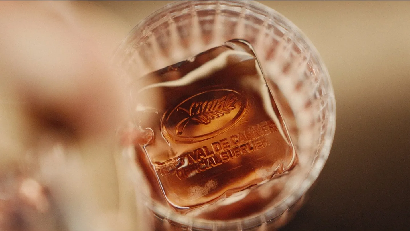 A close-up view of a glass with a brown drink and a single large ice cube engraved with the Cannes Film Festival’s palm logo and text reading FESTIVAL DE CANNES OFFICIAL SUPPLIER.