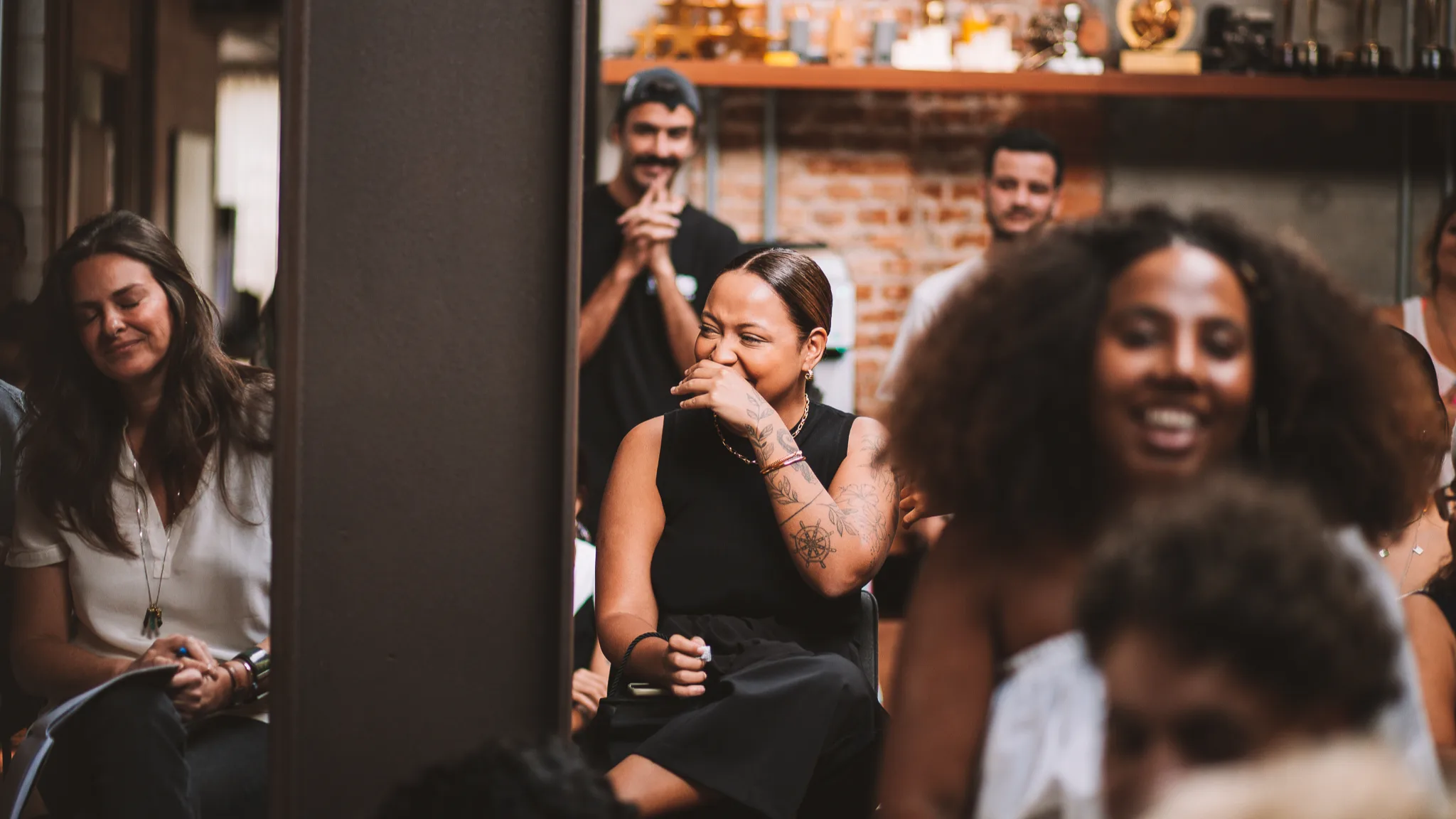 A group of people sit close together indoors, laughing and smiling during what appears to be a lively event or gathering. The focus is on a woman in black, covering her mouth while smiling.