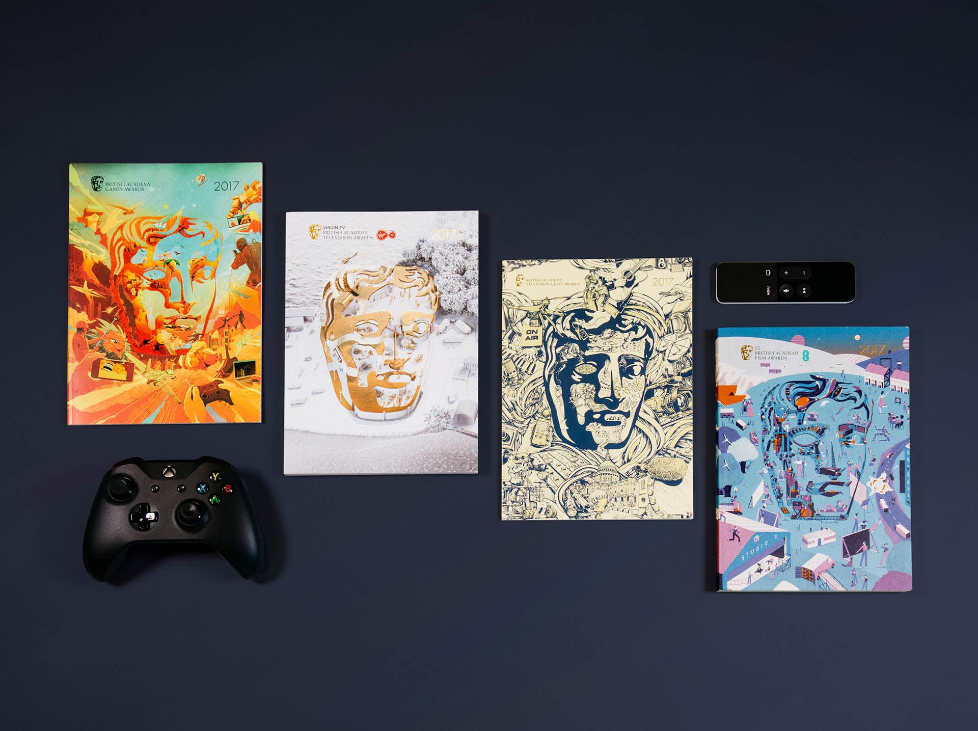 A black game controller and a remote are arranged next to four BAFTA Game Awards booklets with stylized masks and vibrant, artistic covers on a dark background.