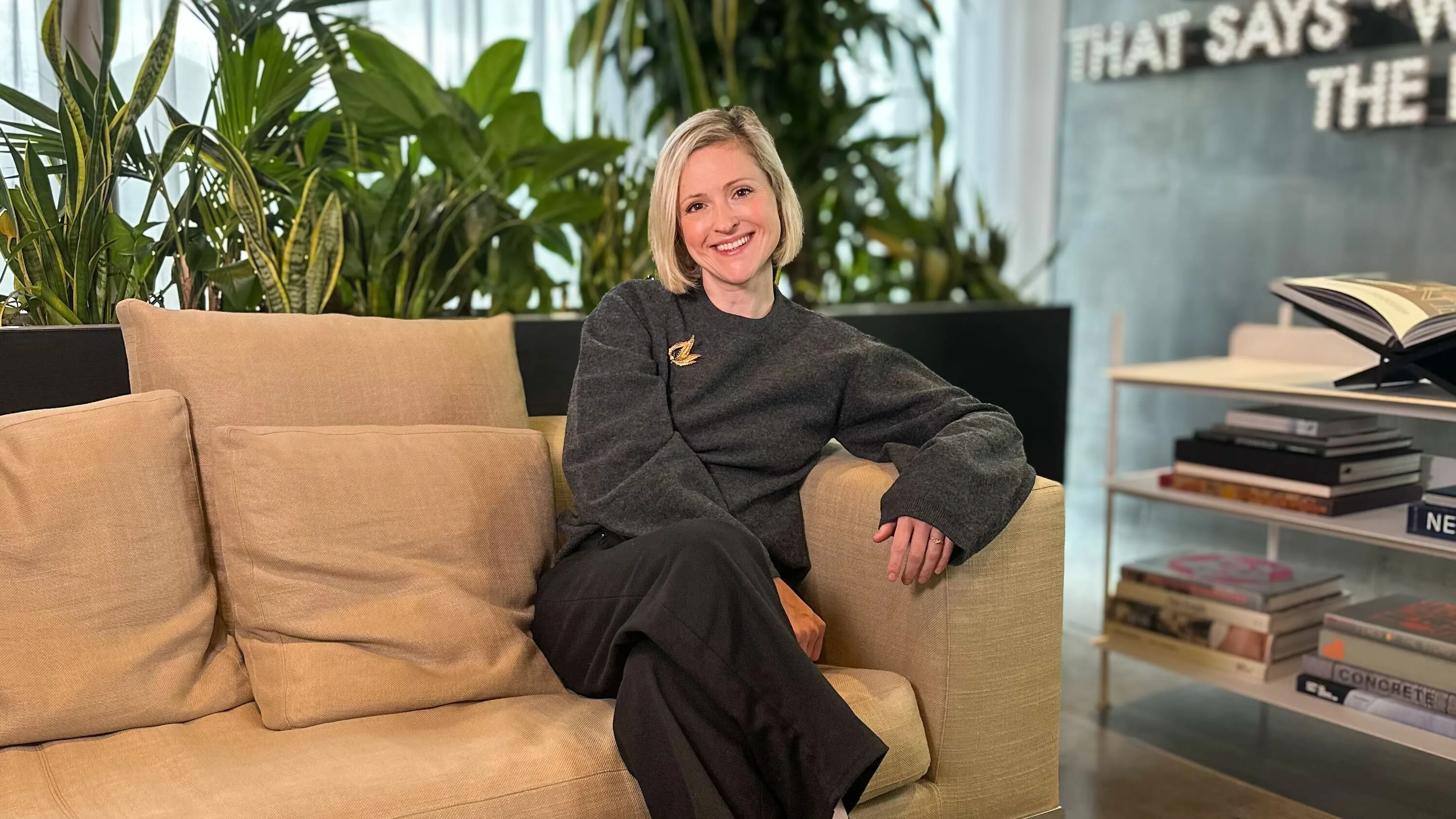 A woman with short blonde hair sits on a beige couch, smiling. She wears a dark sweater and black pants. There are green plants behind her and a bookshelf with books on the right. The setting looks modern and bright.