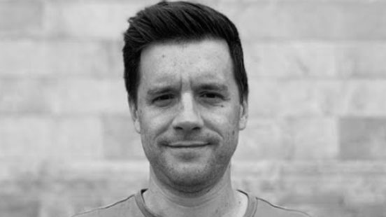 A man with short, dark hair is standing outside in front of a light-colored stone wall, looking directly at the camera with a neutral expression. The image is in black and white.