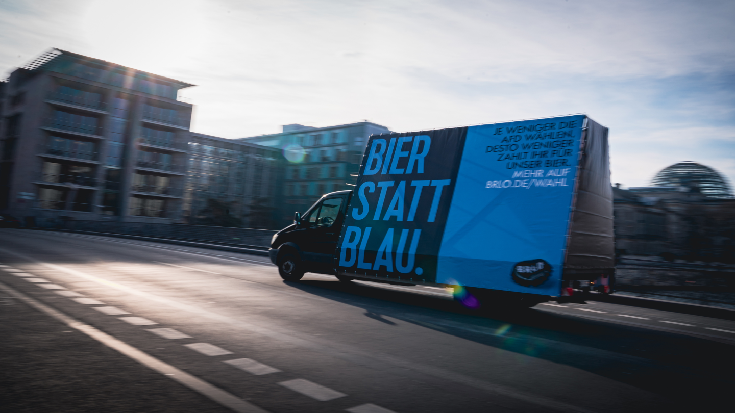 A truck with a billboard that reads Bier statt Blau. drives down a city street. The sign also includes German text and is partially sunlit, with modern buildings and a glass dome in the background.