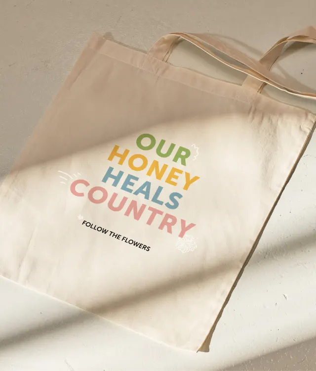 A beige tote bag with the words OUR HONEY HEALS COUNTRY in colorful letters and FOLLOW THE FLOWERS in black text, lying on a light surface in natural sunlight.