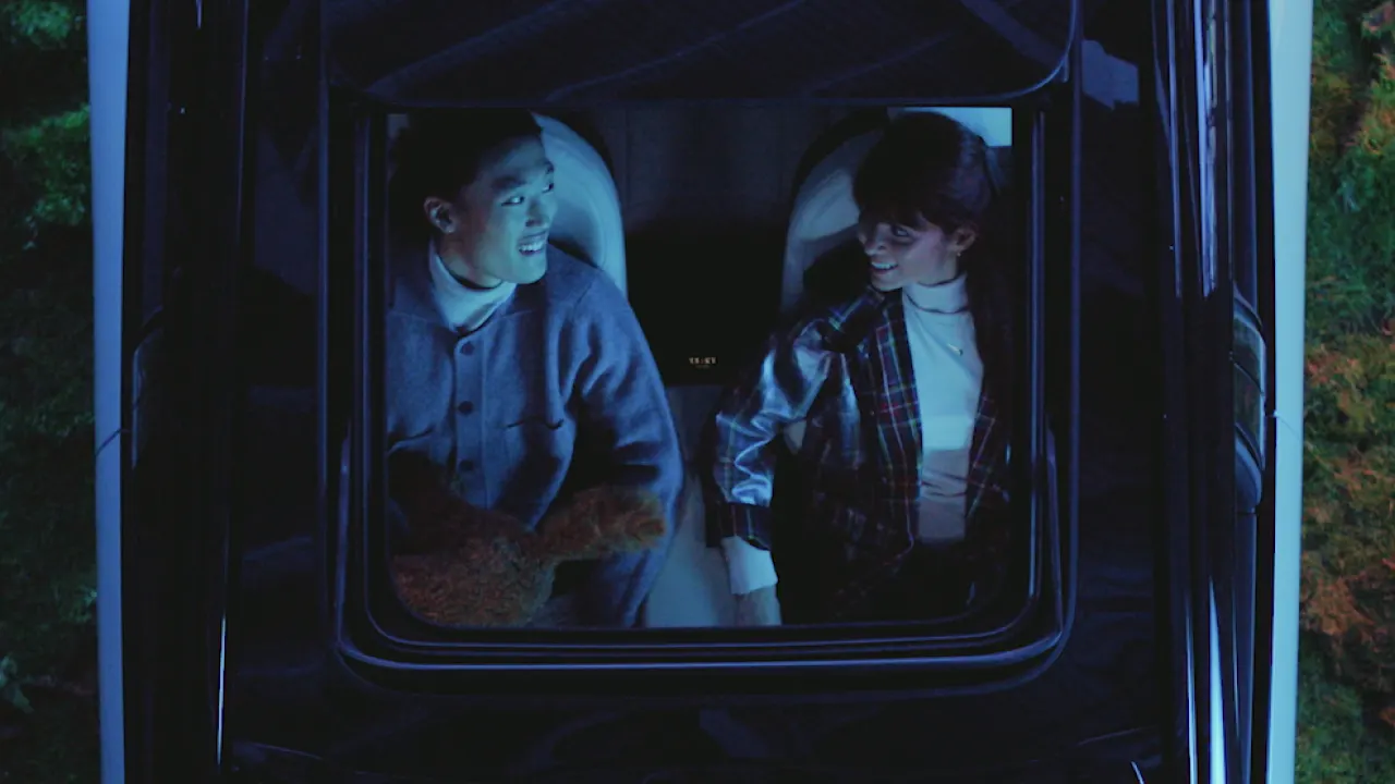 A man and a woman sit side by side in a car, viewed from above through the sunroof at night, smiling at each other under blue lighting.