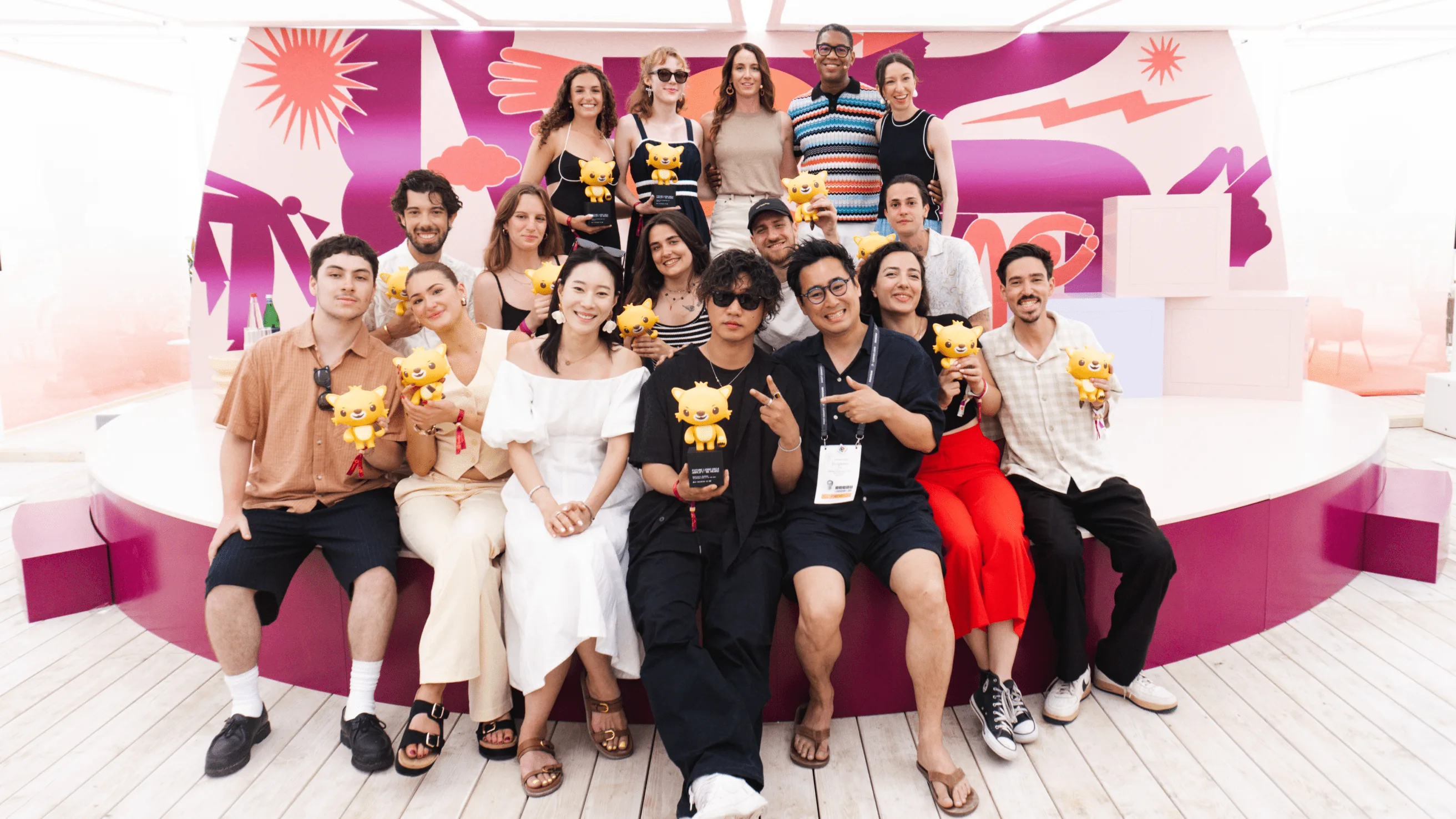 A group of people pose together, smiling, holding yellow cartoon lion trophies. They are on a modern white and purple stage with colorful abstract art in the background.