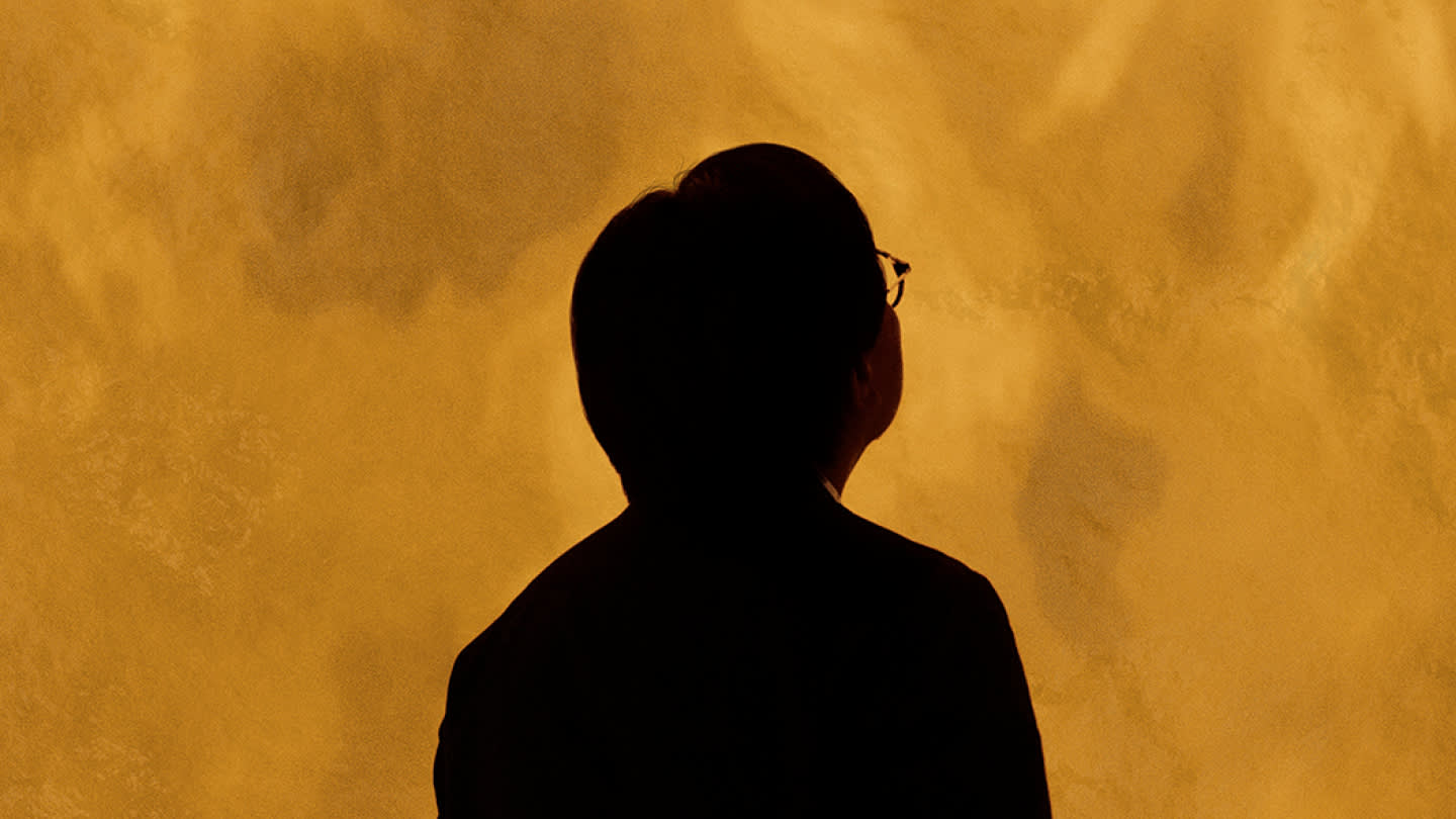 A person with short dark hair, wearing glasses and a dark top, is seen from behind, looking up at a bright, textured golden-yellow background.