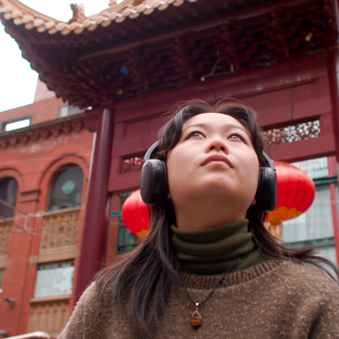 A person wearing headphones and a brown sweater looks upward in front of a traditional Chinese gate with red lanterns hanging behind them.