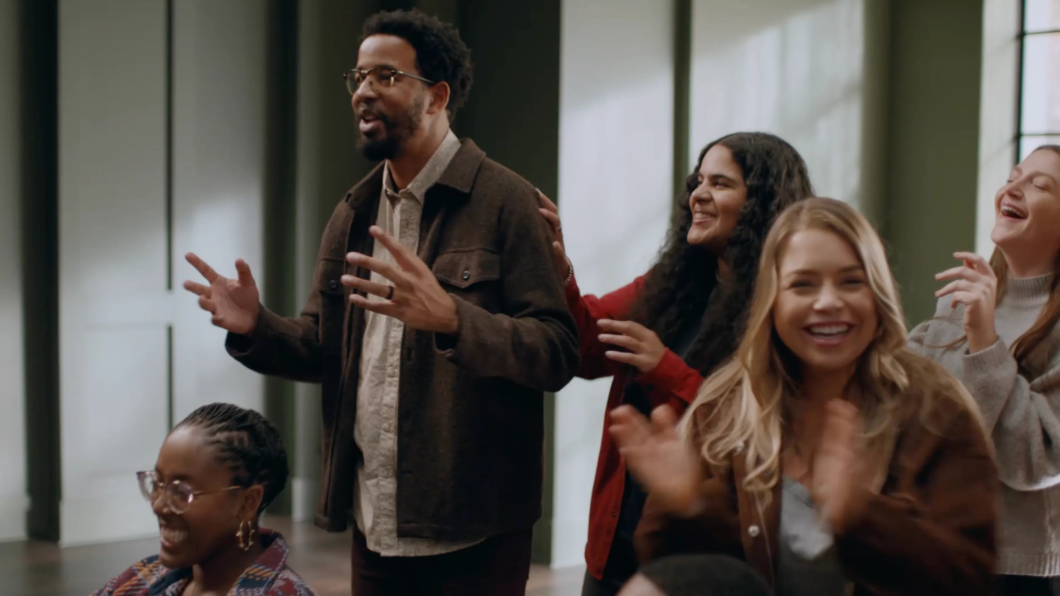 A group of people indoors, smiling, clapping, and standing close together. One man stands while others sit or stand nearby, all appearing happy and engaged in a lively moment.