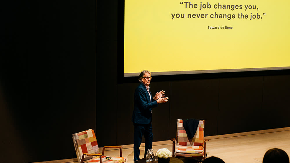 A man stands onstage beside two colorful chairs, speaking in front of a yellow screen displaying the quote: “The job changes you, you never change the job.” – Edward de Bono.