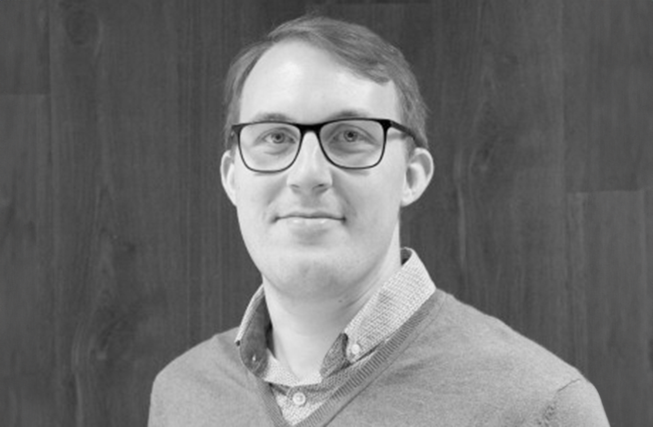A person with short hair and glasses, wearing a collared shirt and sweater, stands in front of a dark wooden background. The image is in black and white.