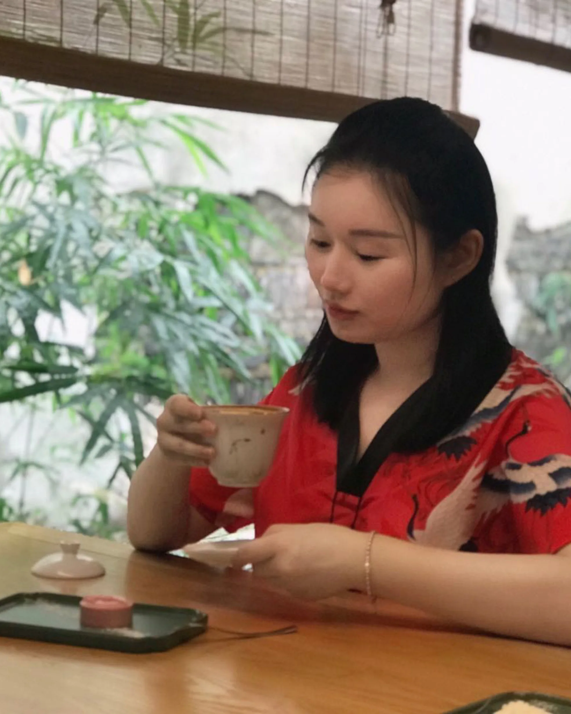 A young woman in a red shirt sits at a wooden table, holding a teacup and looking down. There is a small plate with a pink pastry in front of her, and greenery is visible through the window behind her.