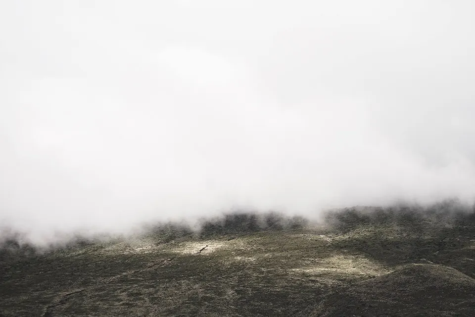 Low, white clouds or mist hover over a rugged, grassy landscape with gentle hills. The scene appears serene and atmospheric, with muted colors and limited visibility due to the thick cloud cover.