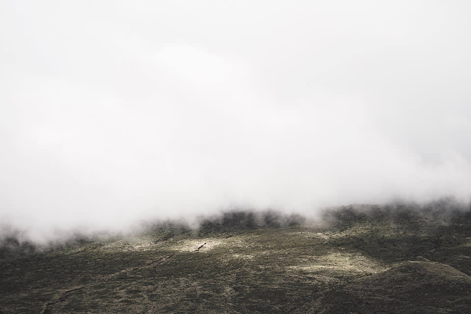 Low, white clouds or mist hover over a rugged, grassy landscape with gentle hills. The scene appears serene and atmospheric, with muted colors and limited visibility due to the thick cloud cover.