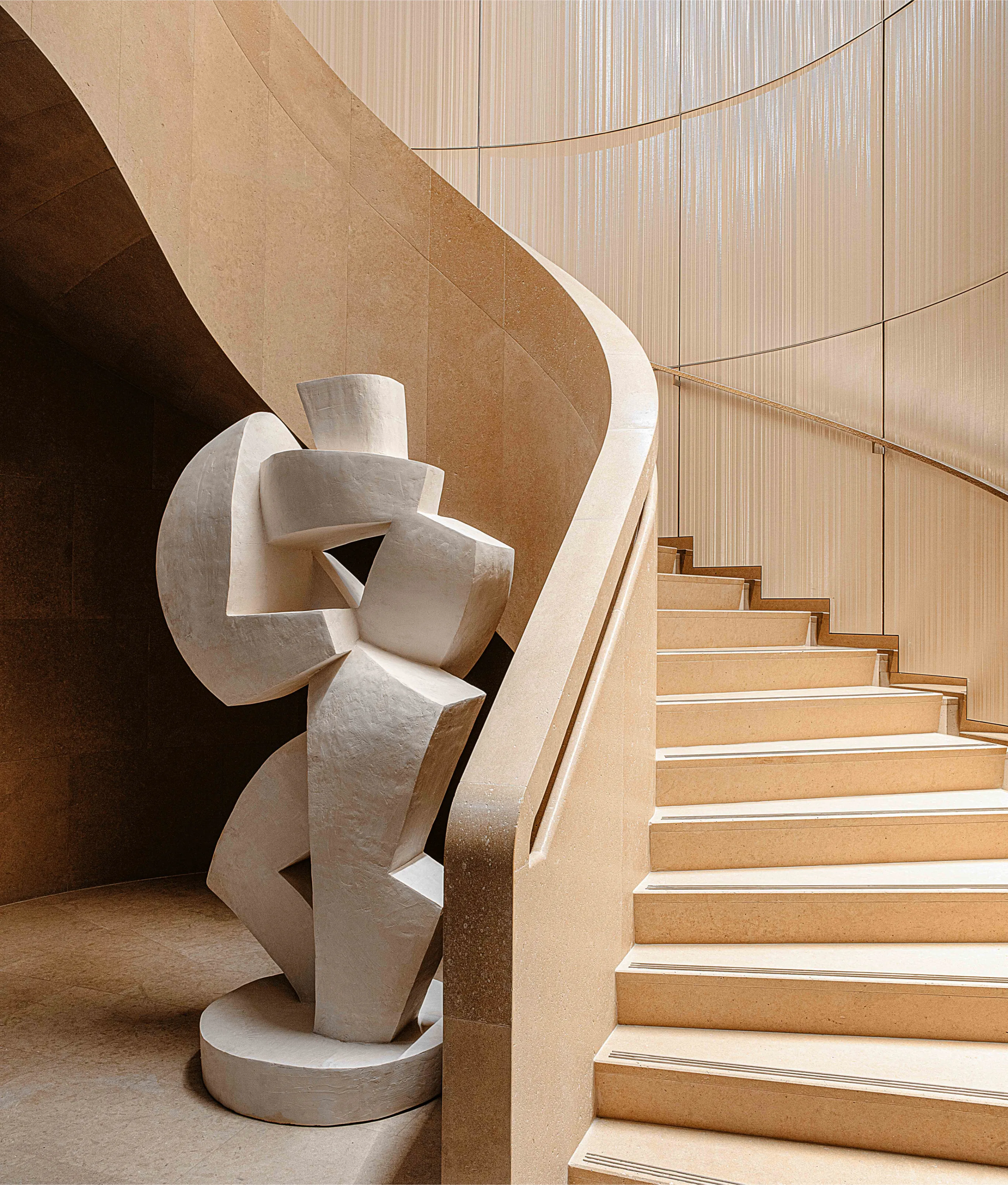 A large abstract white sculpture stands at the foot of a curved beige staircase with smooth, flowing lines, set against a backdrop of vertical textured panels.