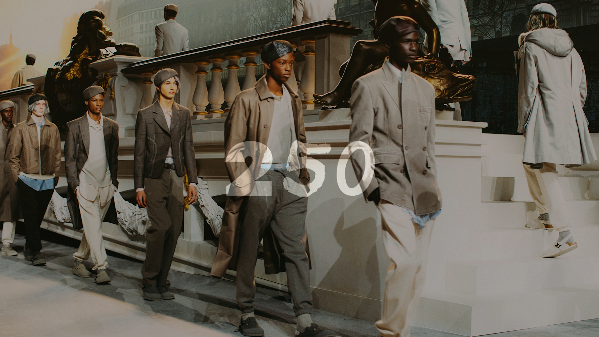 Male models wearing neutral-toned, layered outfits and berets walk down a fashion runway, passing ornate white sculptures and stairs, with a cityscape backdrop and warm lighting.