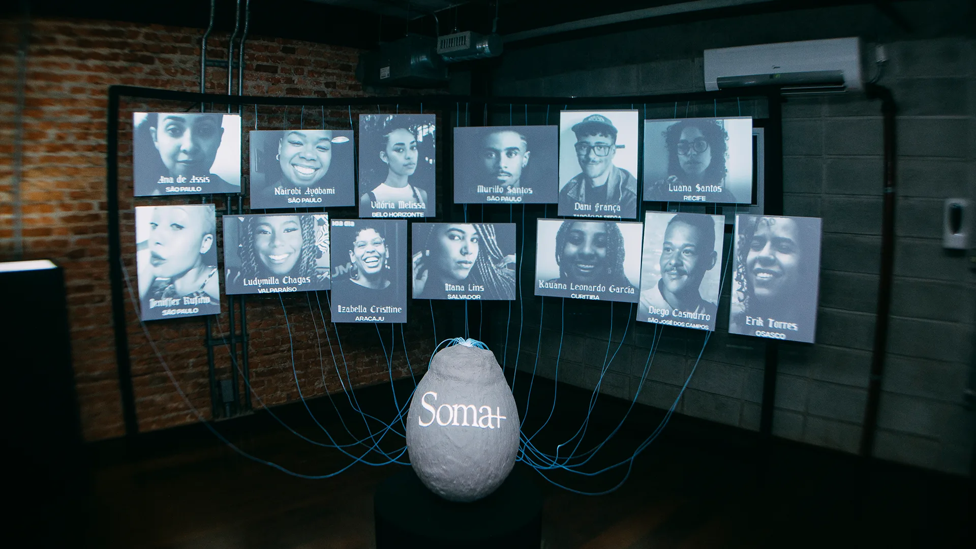 A stone labeled Somar is connected by blue-lit wires to multiple screens displaying black-and-white portraits and names of various individuals in a dimly lit, industrial room with brick walls.
