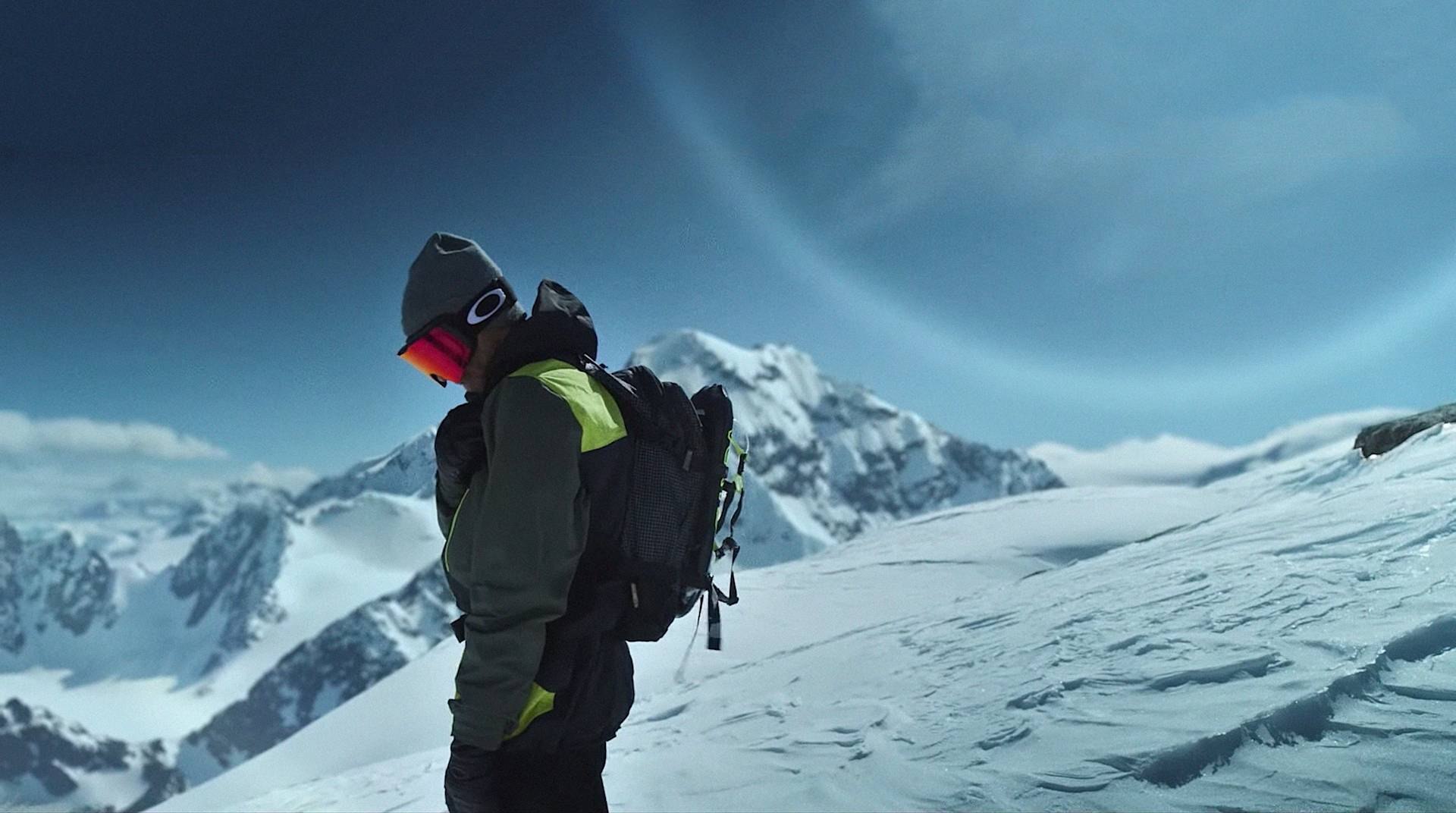 A person wearing winter gear and a backpack stands on a snowy mountain slope, surrounded by snow-covered peaks under a blue sky with a large planet visible in the background.