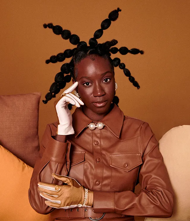 A stylish woman with an intricate, spiky braided hairstyle sits on a sofa, wearing a brown leather shirt, pearl necklace, and two gloves—one white, one tan—against a brown background with neutral-toned cushions.