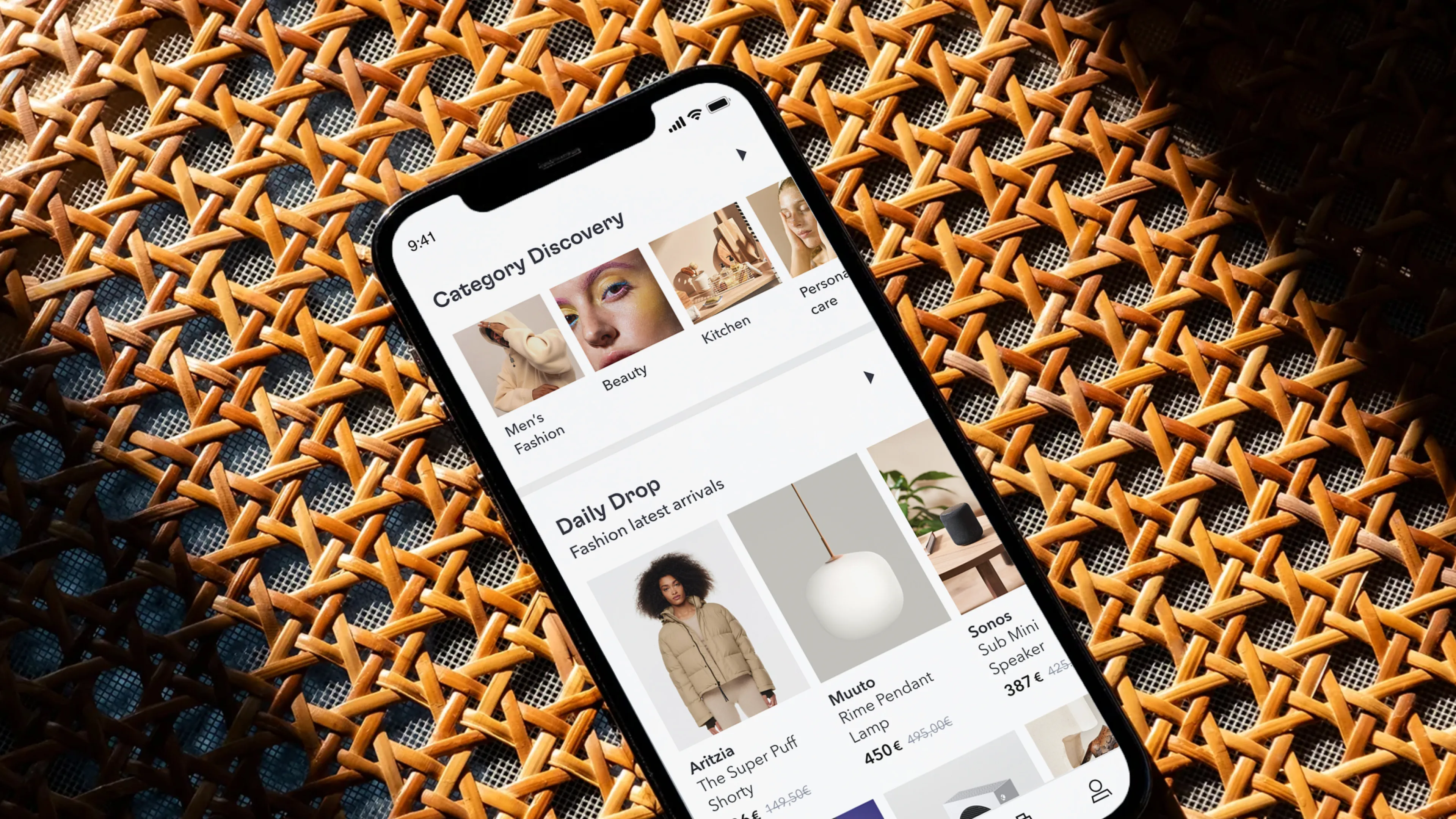 A smartphone displaying a shopping app screen, featuring categories like fashion, beauty, and kitchen, and showcasing daily drop deals, placed on a patterned wicker surface under natural light.