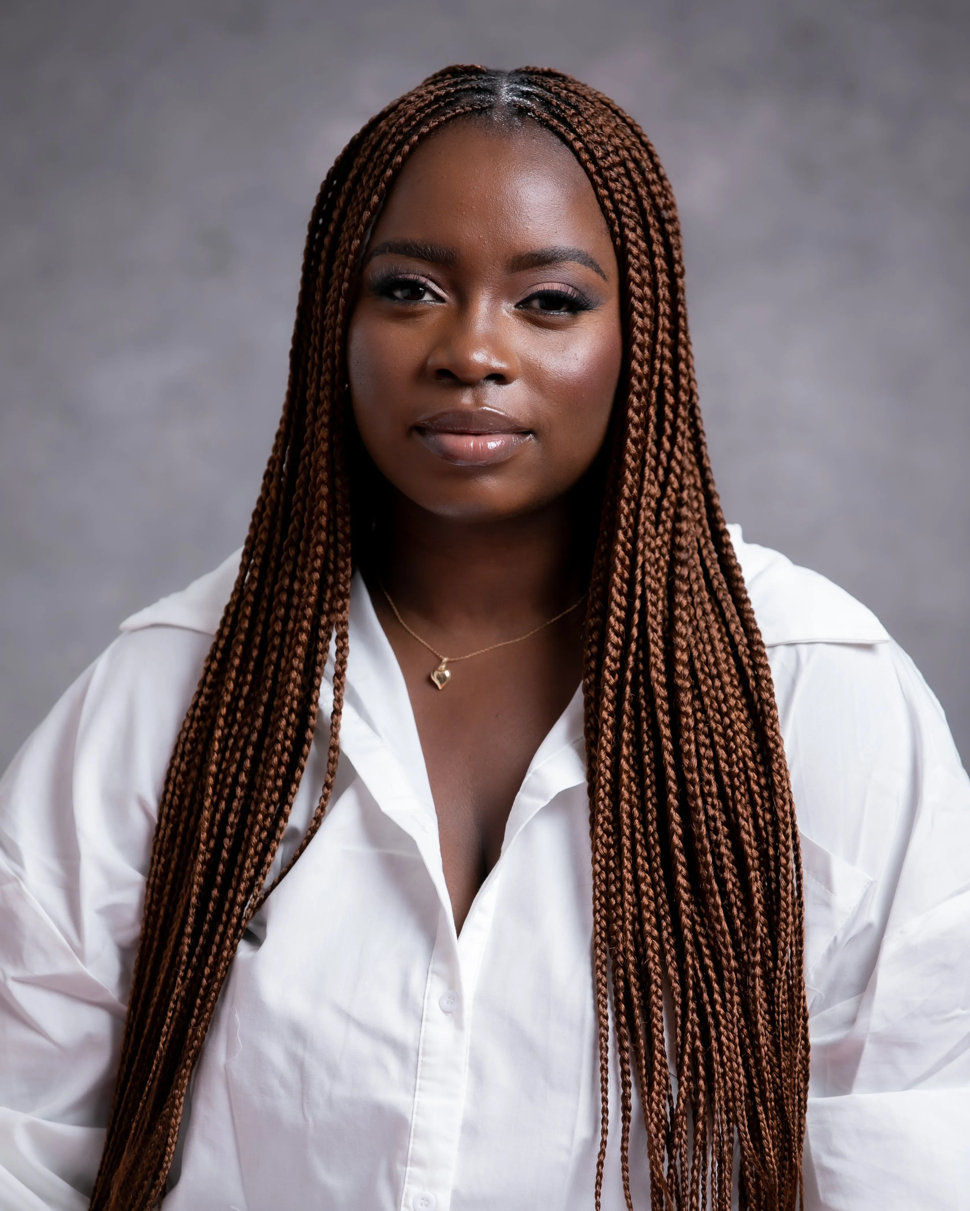 A woman with long, brown box braids wears a white shirt and a simple gold necklace. She is looking at the camera with a neutral expression, posed against a gray background.
