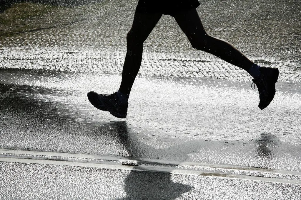 A person in dark athletic clothing runs through a wet street, their feet splashing water as they move. Their shadow and the glistening wet pavement are visible below them.