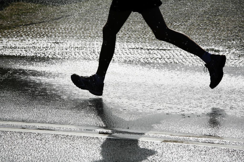 A person in dark athletic clothing runs through a wet street, their feet splashing water as they move. Their shadow and the glistening wet pavement are visible below them.