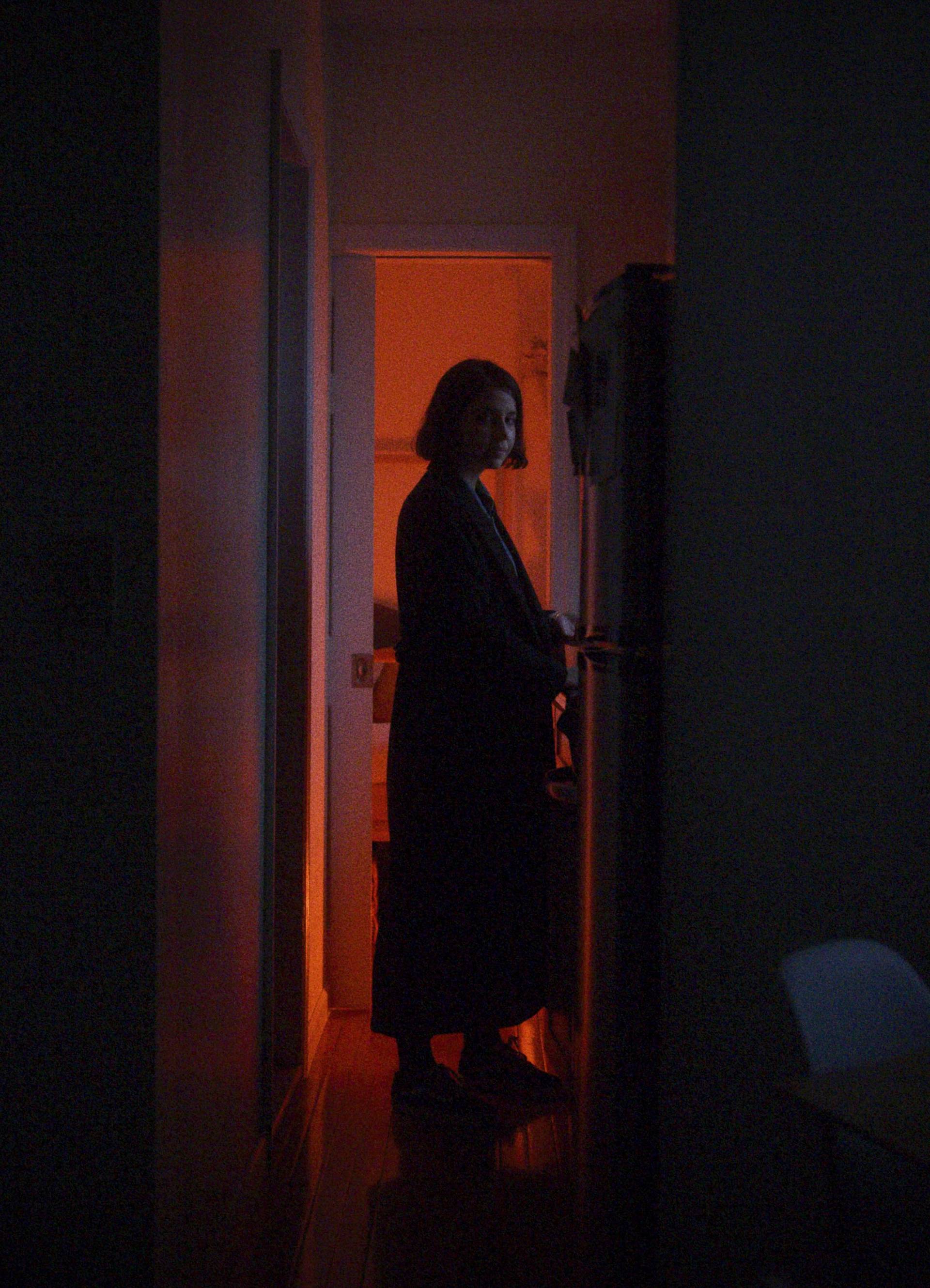 A woman in a dark robe stands by an open refrigerator in a dimly lit hallway. Warm orange light from an adjacent room contrasts with the cool darkness around her.
