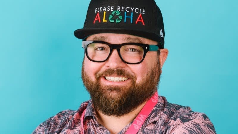 A bearded person wearing glasses, a black cap that says PLEASE RECYCLE ALOHA with a recycling symbol, a pink patterned shirt, and a pink lanyard, smiling in front of a blue background.